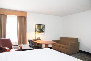 Una habitación de hotel con cama, escritorio, silla, sofá, lámpara y un cuadro en la pared.