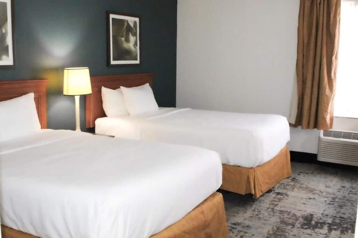 A hotel room with two beds, a nightstand with a lamp, a wall-mounted air conditioner, and a window with curtains.