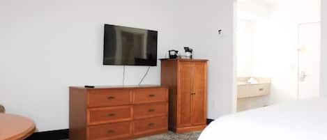 A bedroom with a bed, a wooden dresser, a television mounted on the wall, and a bathroom visible through an open door.