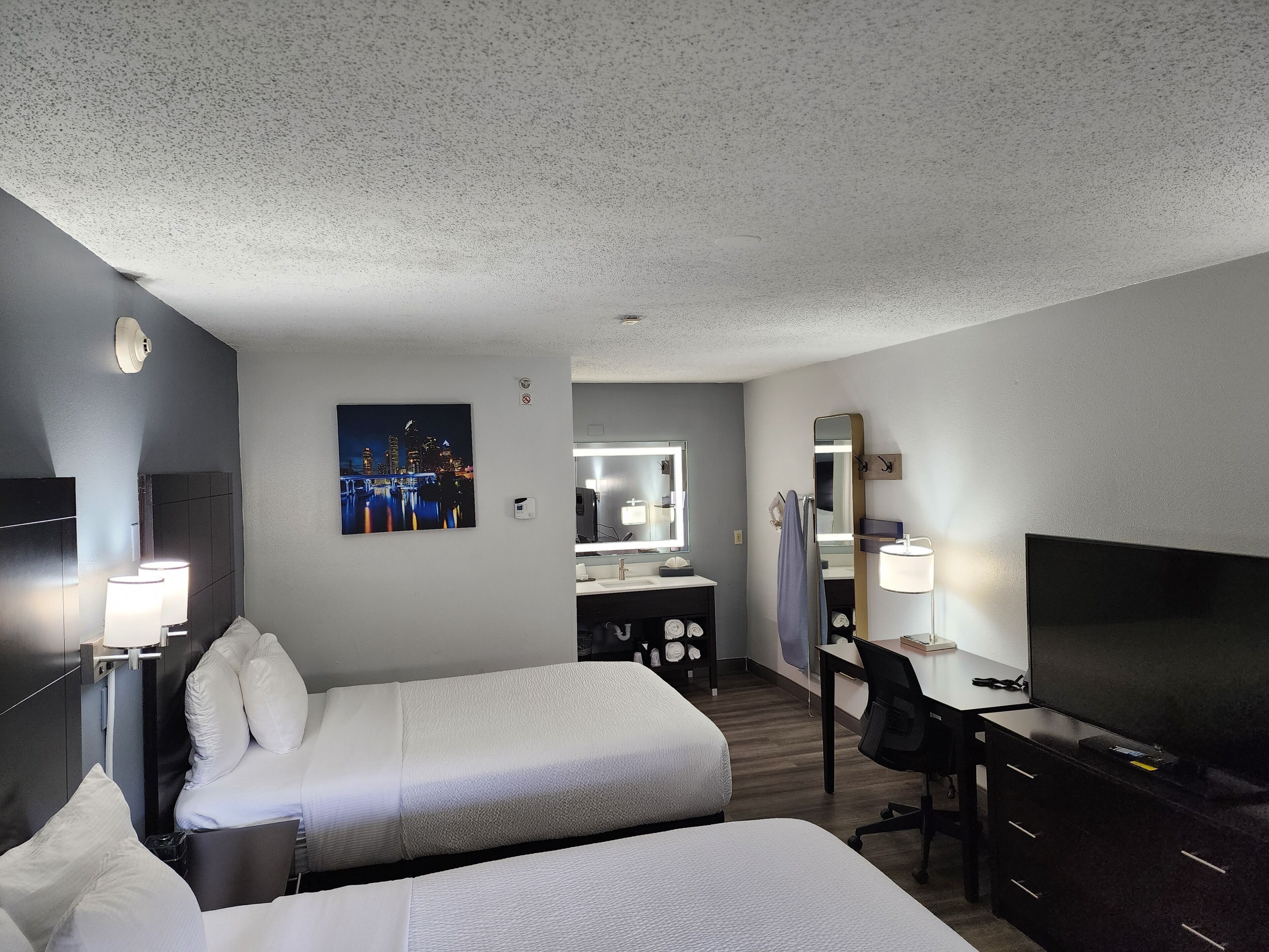 A hotel room with two beds, a desk with a chair, a TV, a mirror, a bathroom with a sink and mirror, and a cityscape painting on the wall.