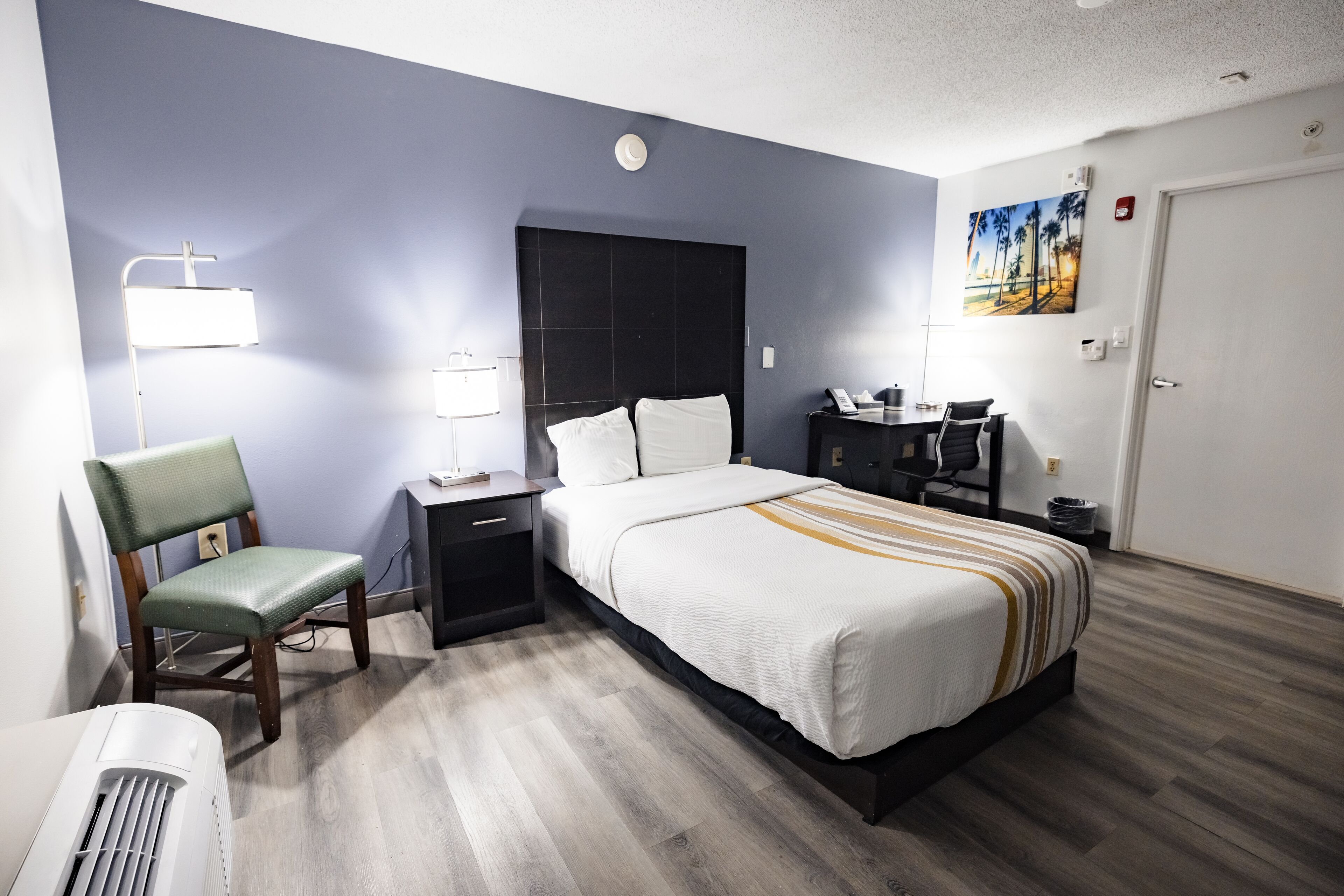 A hotel room with a bed, a desk, a chair, a lamp, and a painting on the wall.