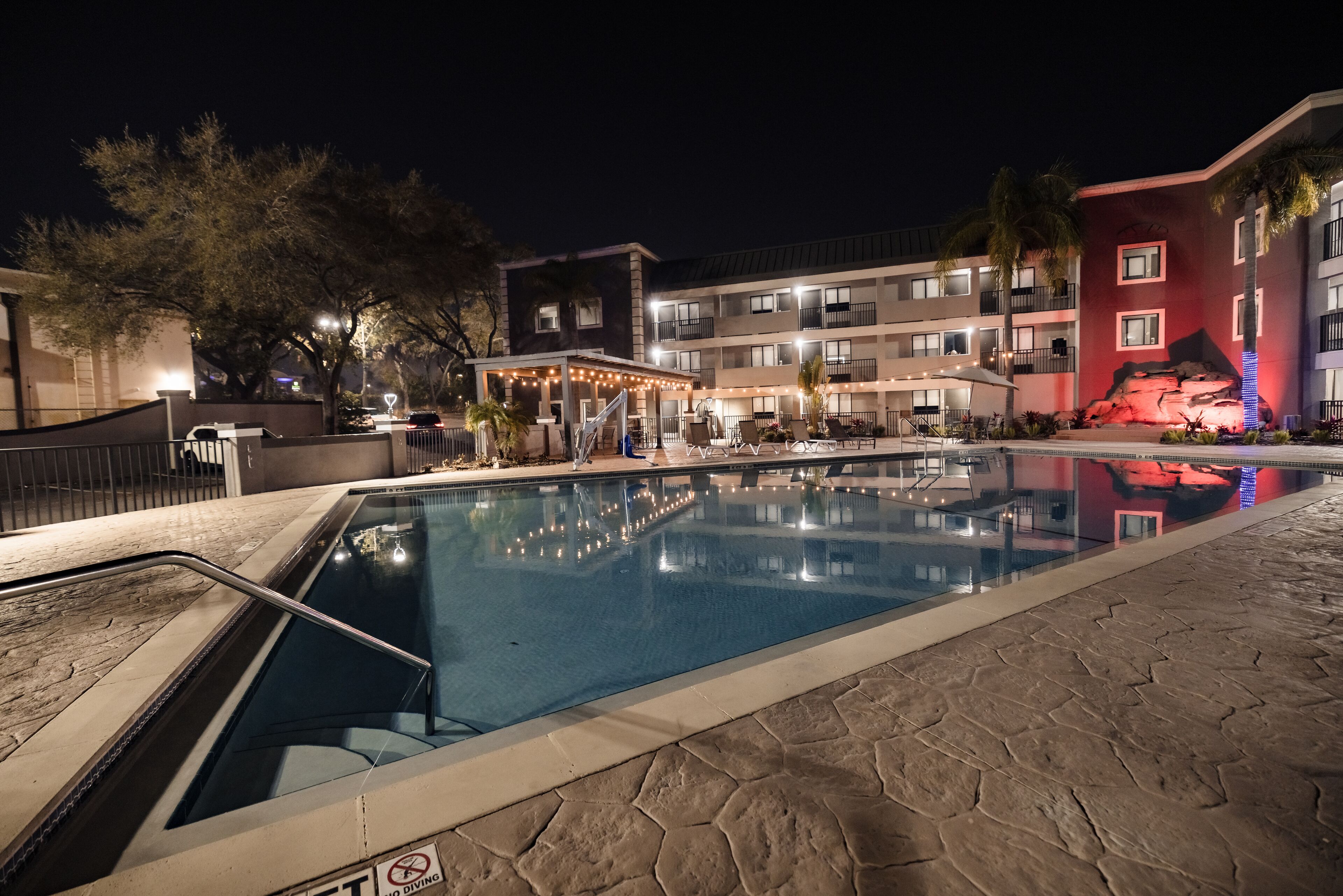 A swimming pool area with a lifeguard stand, a covered seating area, and a building with balconies and a red-lit wall.