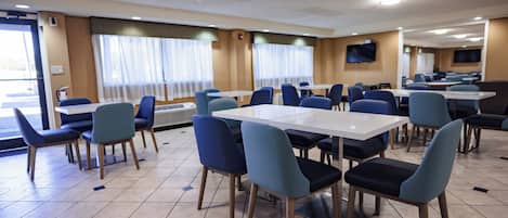 A dining area with tables and chairs, large windows with curtains, and a flat-screen TV on the wall.