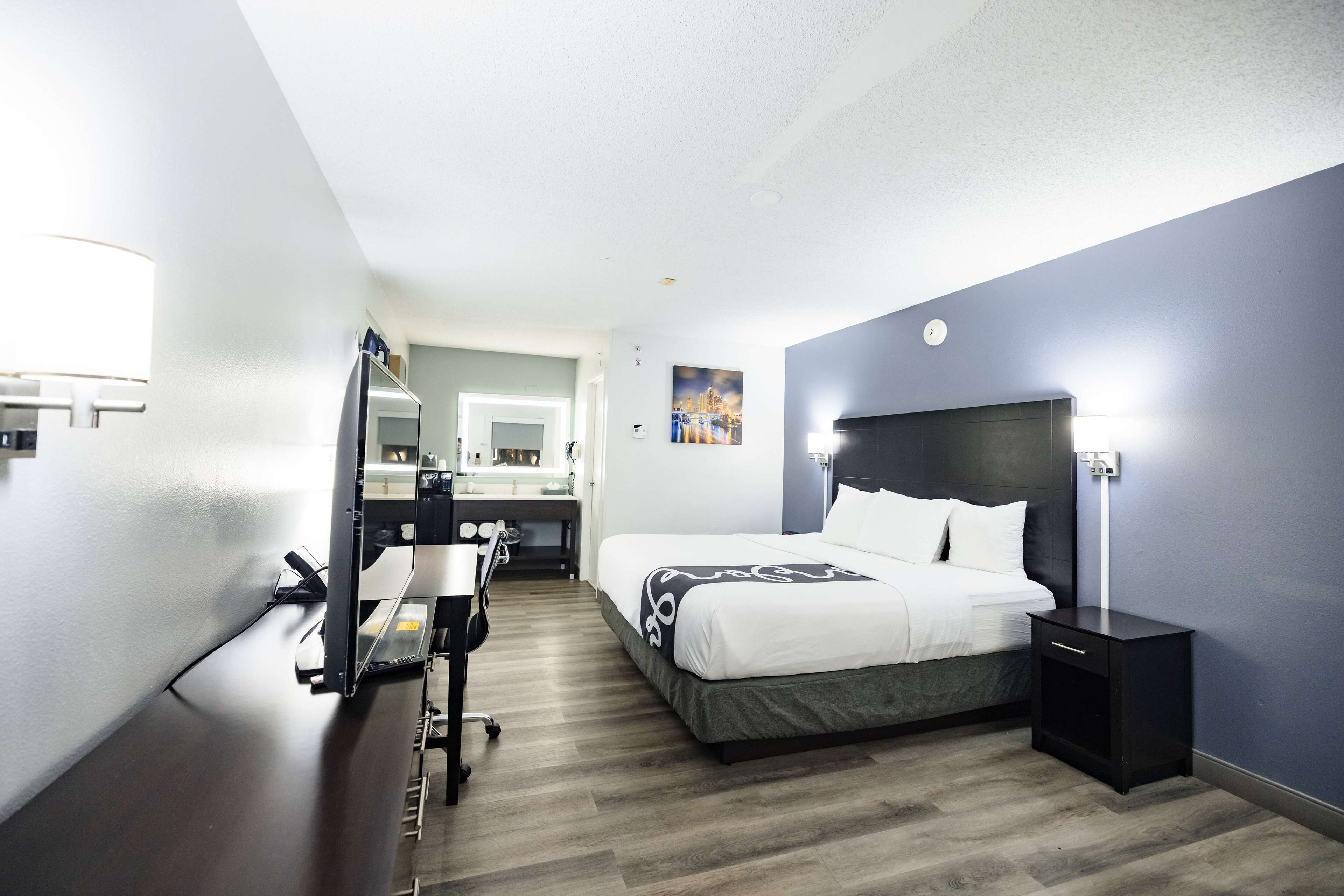 A hotel room with a bed, a desk with a computer, a mirror, and a nightstand.