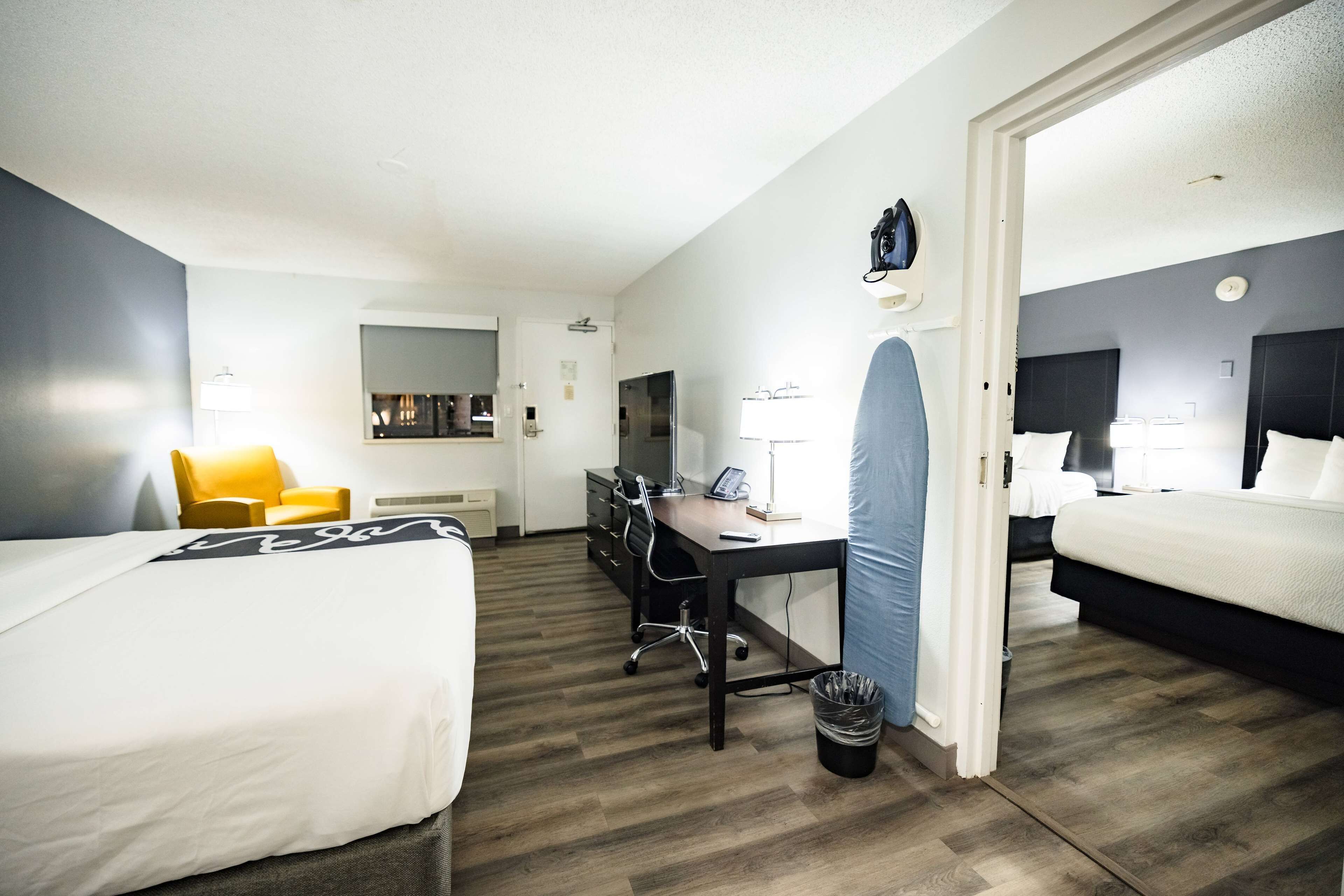 A hotel room with two beds, a desk, a chair, a trash bin, and a door leading to another room.