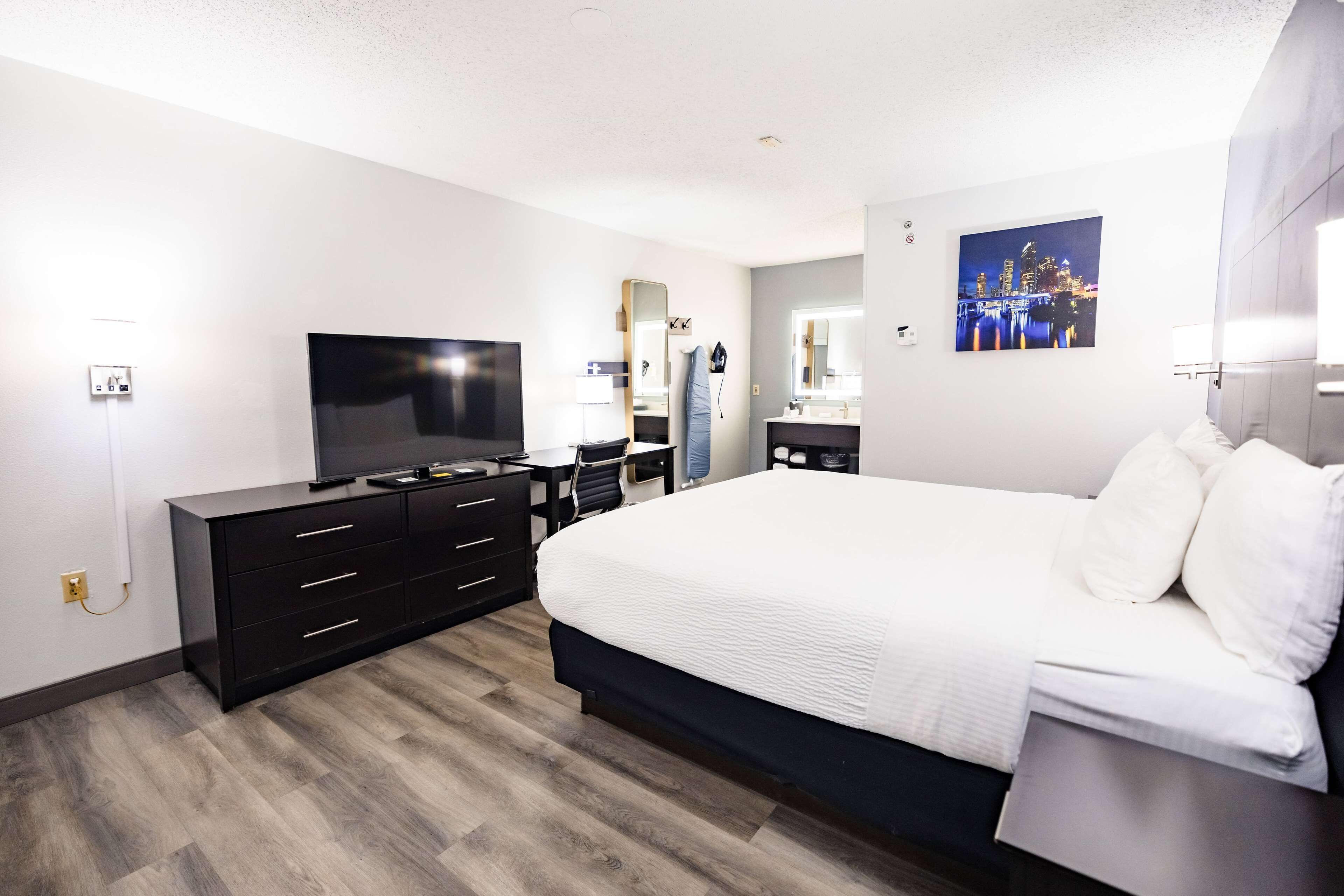 A modern hotel room with a large bed, a flat-screen TV, a desk with a chair, a vanity with a mirror, and a cityscape painting on the wall.