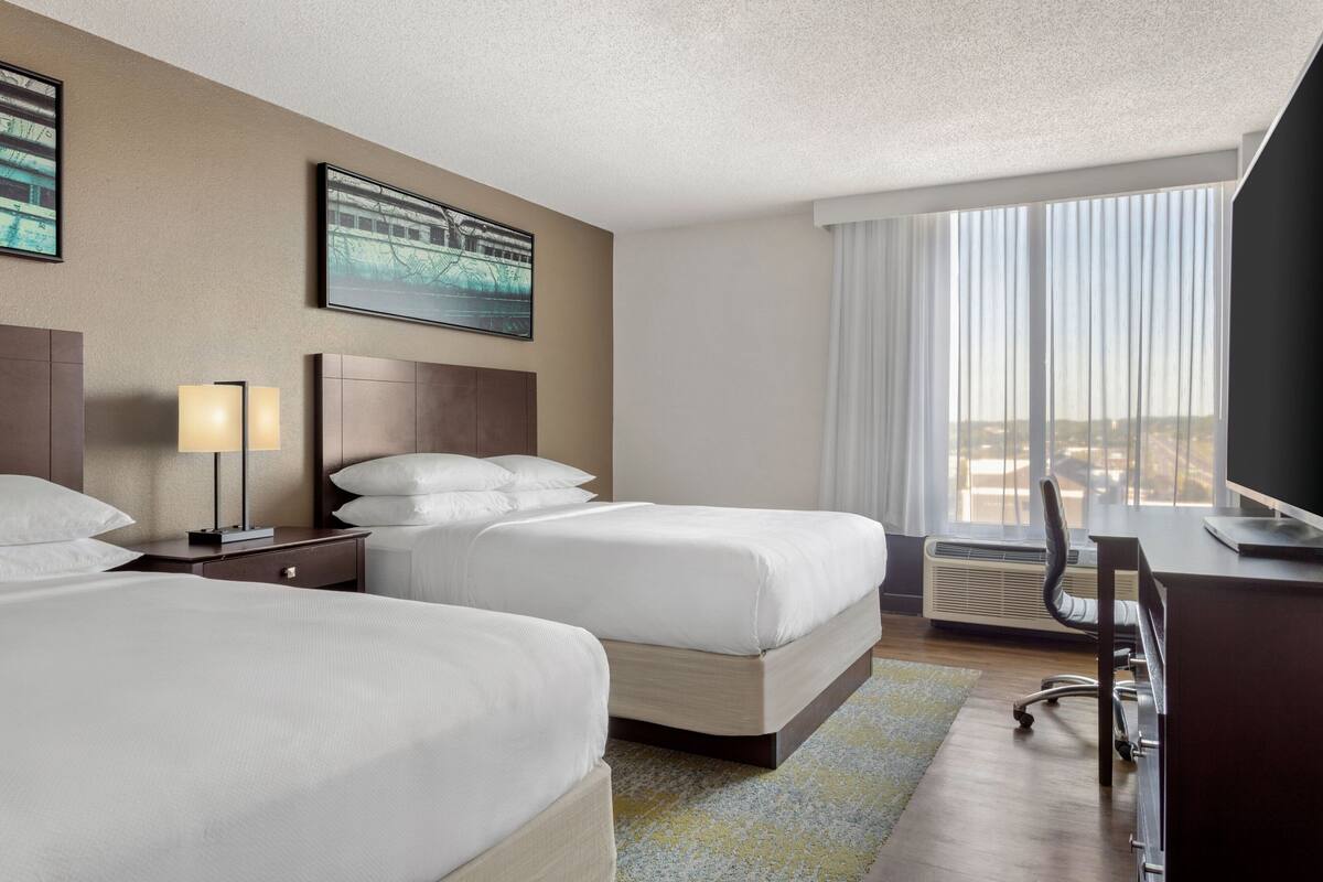 A hotel room with two beds, a desk, a chair, and a large window with curtains.