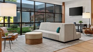 A modern living room with a beige sofa, a round ottoman, and a coffee table. There is a large window with a view of greenery and a building outside. A flat-screen TV is mounted on the wall, and there are two floor lamps. - Delta Hotels by Marriott Bristol (Bristol)