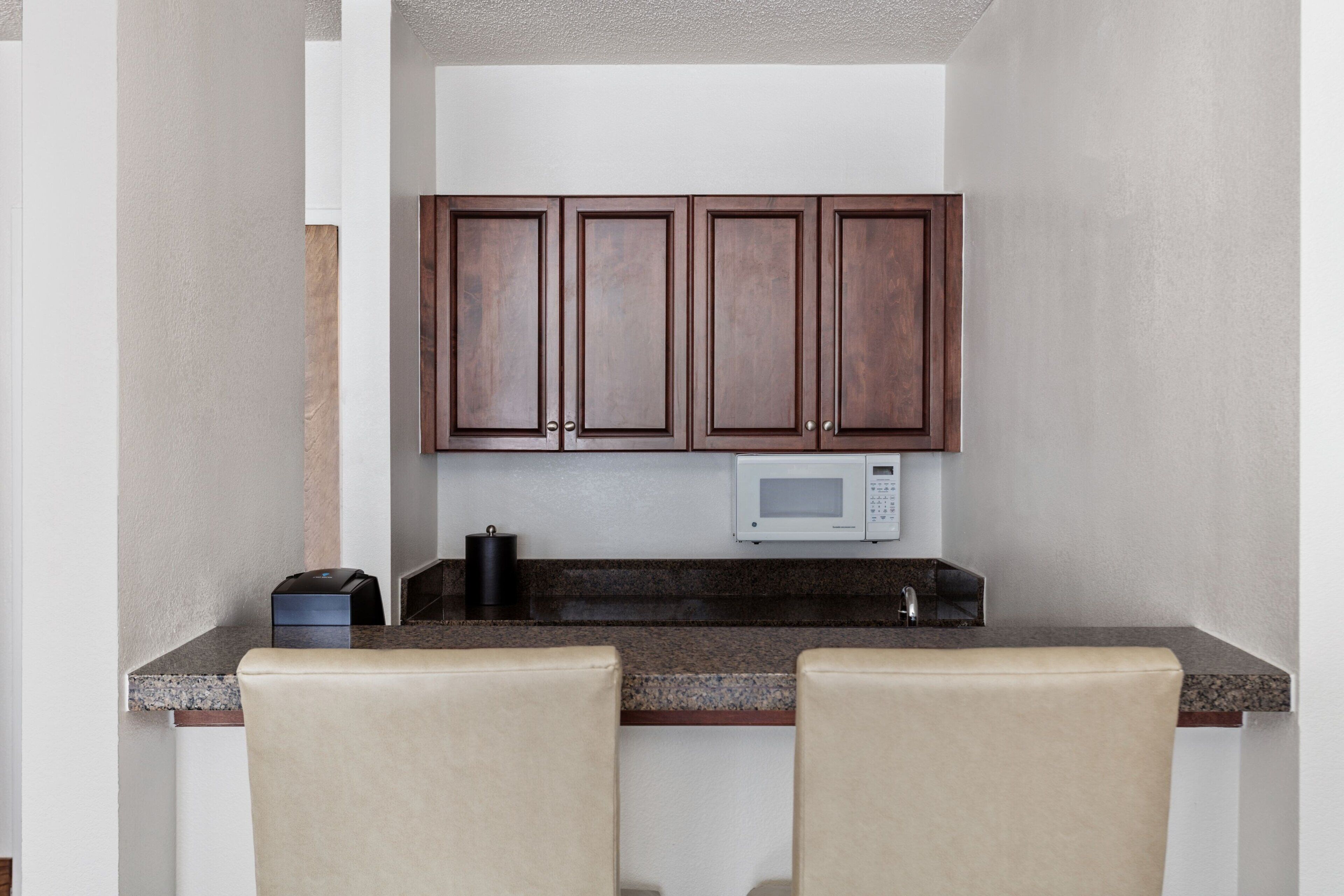 A small kitchen with dark wooden cabinets, a microwave, and a coffee maker on the counter. There are two beige chairs in front of the counter.