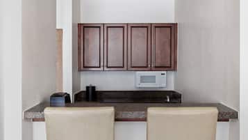 Una pequeña cocina con armarios de madera oscura, un microondas y una cafetera sobre la encimera. Hay dos sillas color beige frente a la encimera.