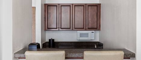 A small kitchen with dark wooden cabinets, a microwave, and a coffee maker on the counter. There are two beige chairs in front of the counter.