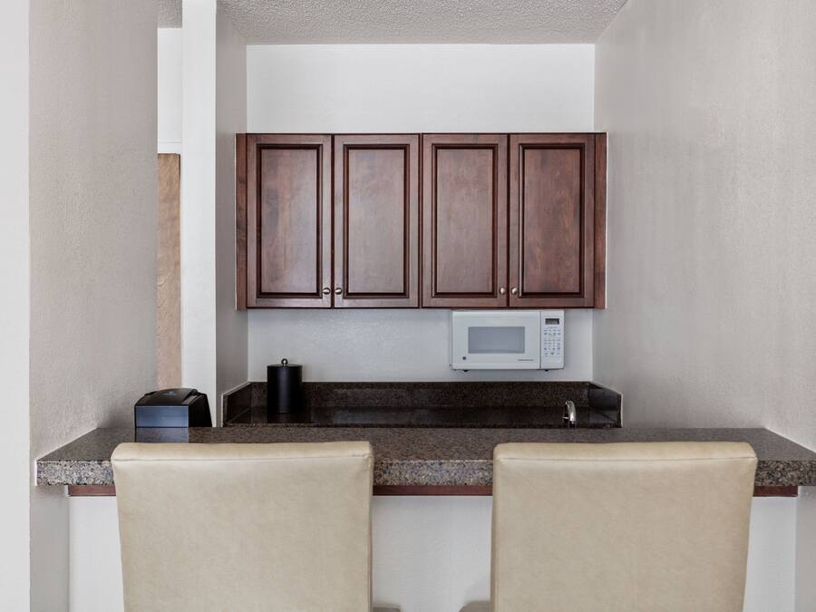 Una pequeña cocina con armarios de madera oscura, un microondas y una cafetera sobre la encimera. Hay dos sillas color beige frente a la encimera.