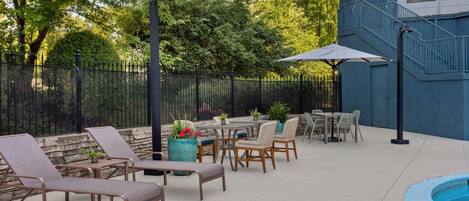 An outdoor patio with lounge chairs, a table, and chairs, surrounded by a pool and a fence.