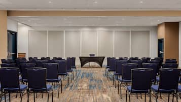 A conference room with rows of chairs facing a table, carpeted floor, and a plain wall with a door on the left.