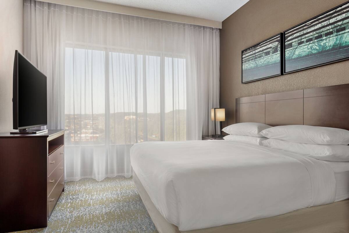 A hotel room with a large bed, a flat-screen TV on a dresser, a window with sheer curtains, and two framed pictures on the wall.