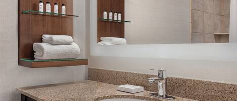 Une salle de bain moderne avec un comptoir en granit, un lavabo blanc et un miroir au-dessus. On y trouve des étagères contenant des articles de toilette et des serviettes.
