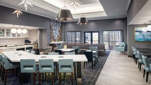 Un hall d’hôtel moderne doté d’un espace de restauration central, d’une cuisine et d’un coin salon offrant une vue sur l’extérieur.
