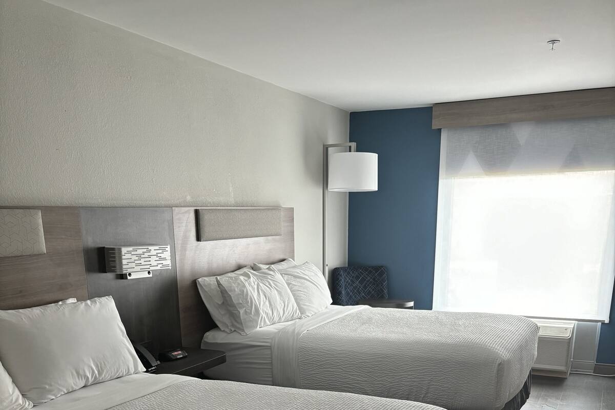 Una habitación de hotel con dos camas, una mesita de noche, una lámpara y un ventanal con cortinas.