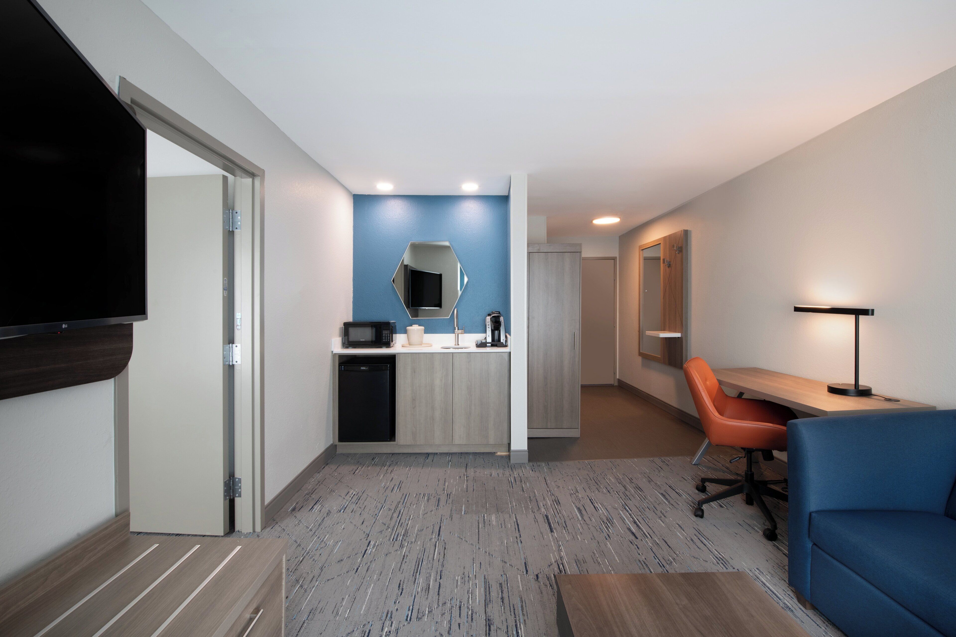 A modern hotel room with a kitchenette, a flat-screen TV, a desk with a chair, and a blue sofa.