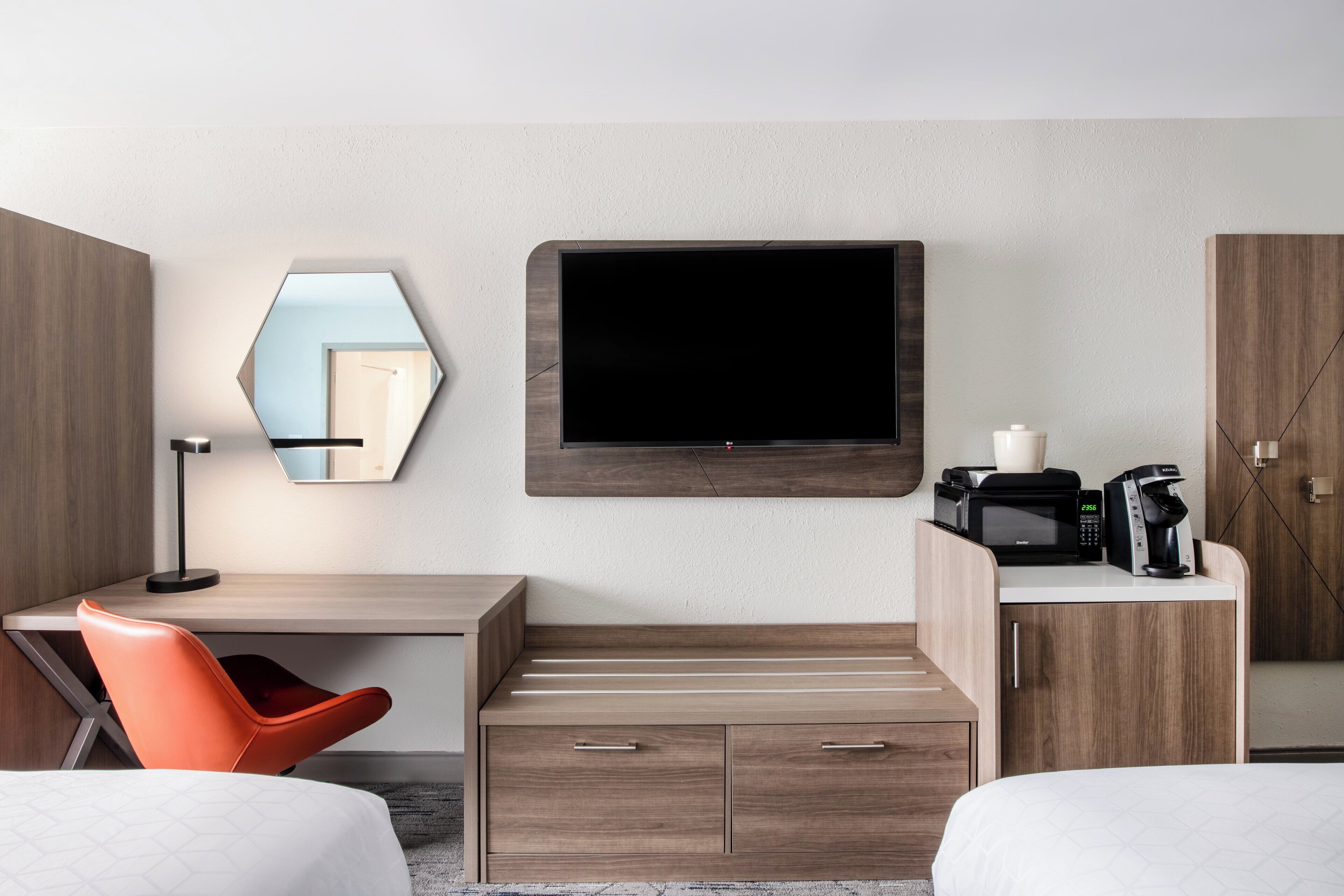 A hotel room with a wooden desk, orange chair, a flat-screen TV mounted on the wall, a microwave, and a coffee maker.