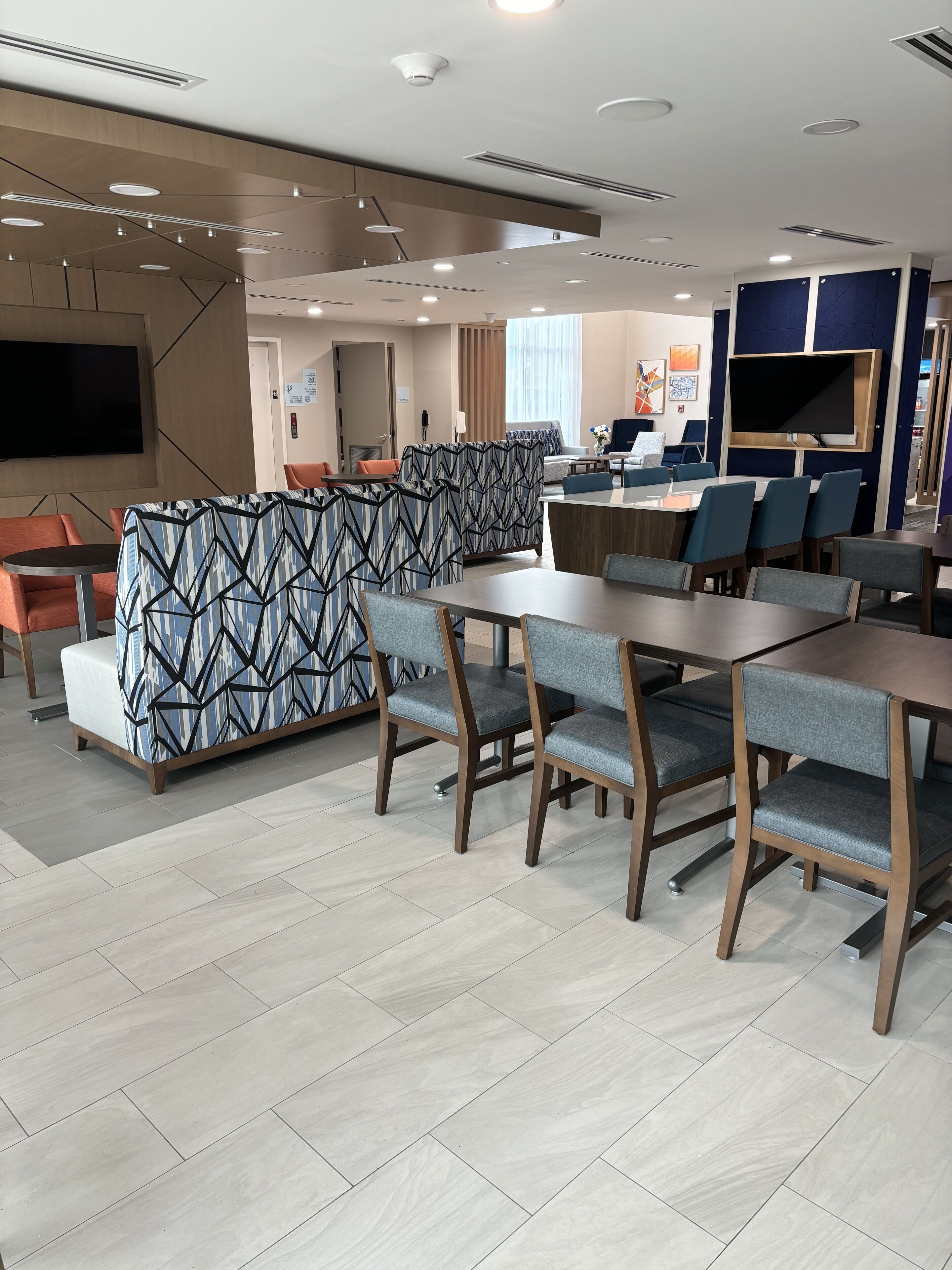 A modern hotel lobby with a seating area featuring patterned sofas and a dining area with wooden tables and chairs.