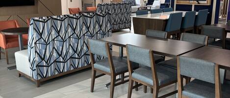 A modern hotel lobby with a seating area featuring patterned sofas and a dining area with wooden tables and chairs.