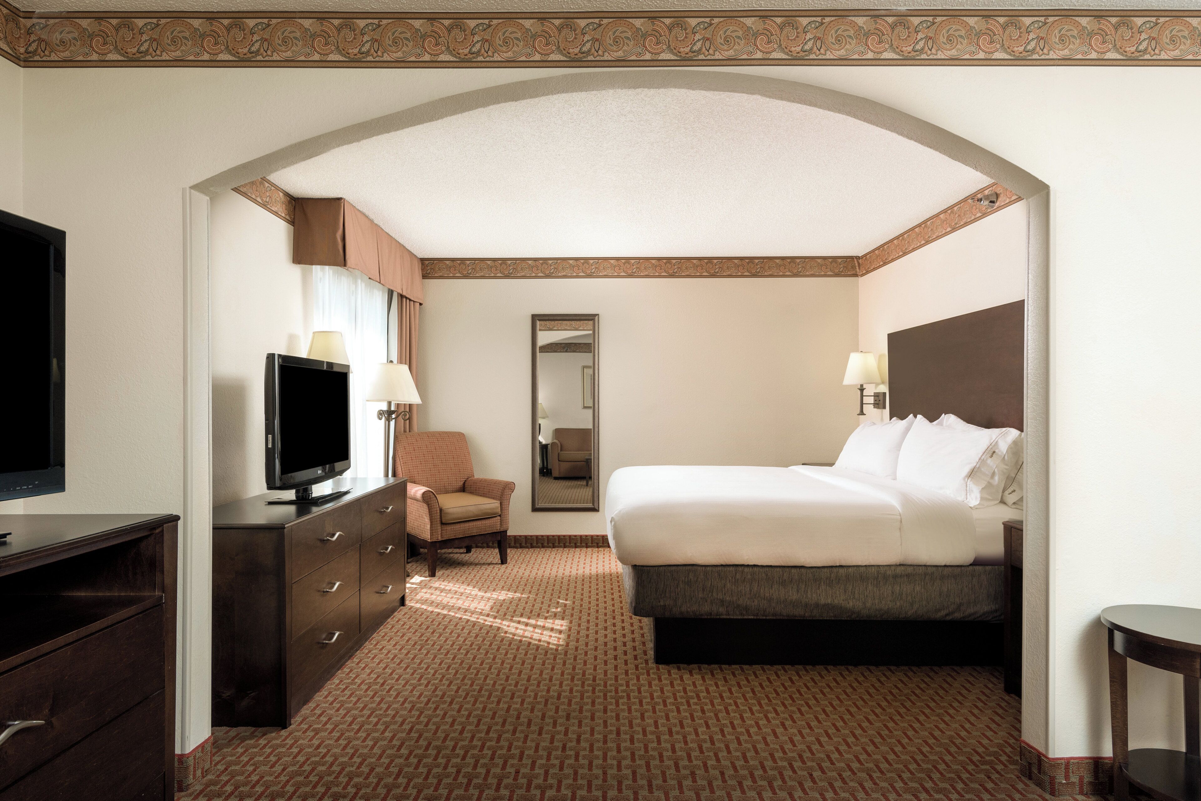 A hotel room with a bed, a television, a chair, a dresser, and a mirror.