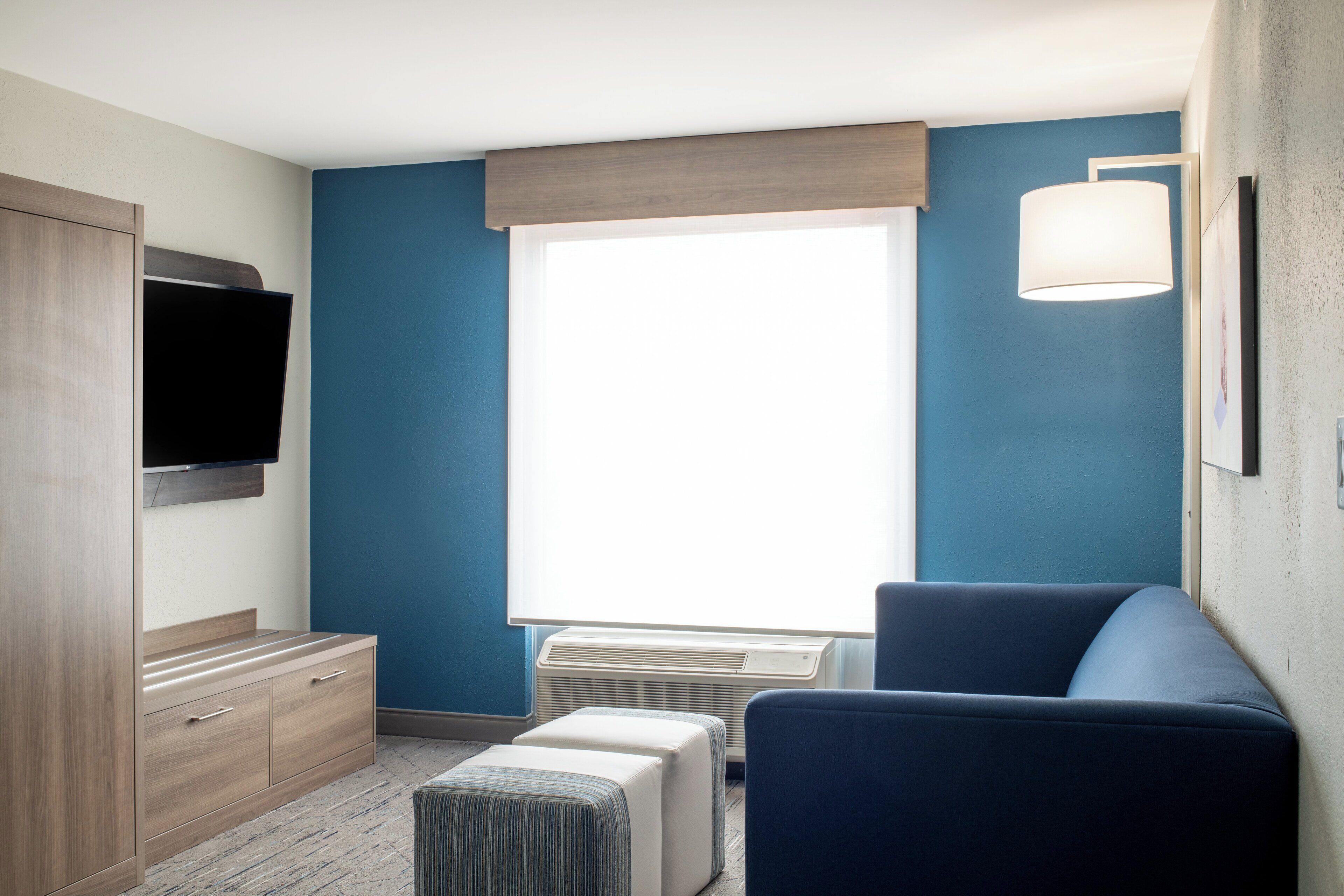 A hotel room with a large window, a flat-screen TV mounted on the wall, a sofa, a small ottoman, and a wooden cabinet.
