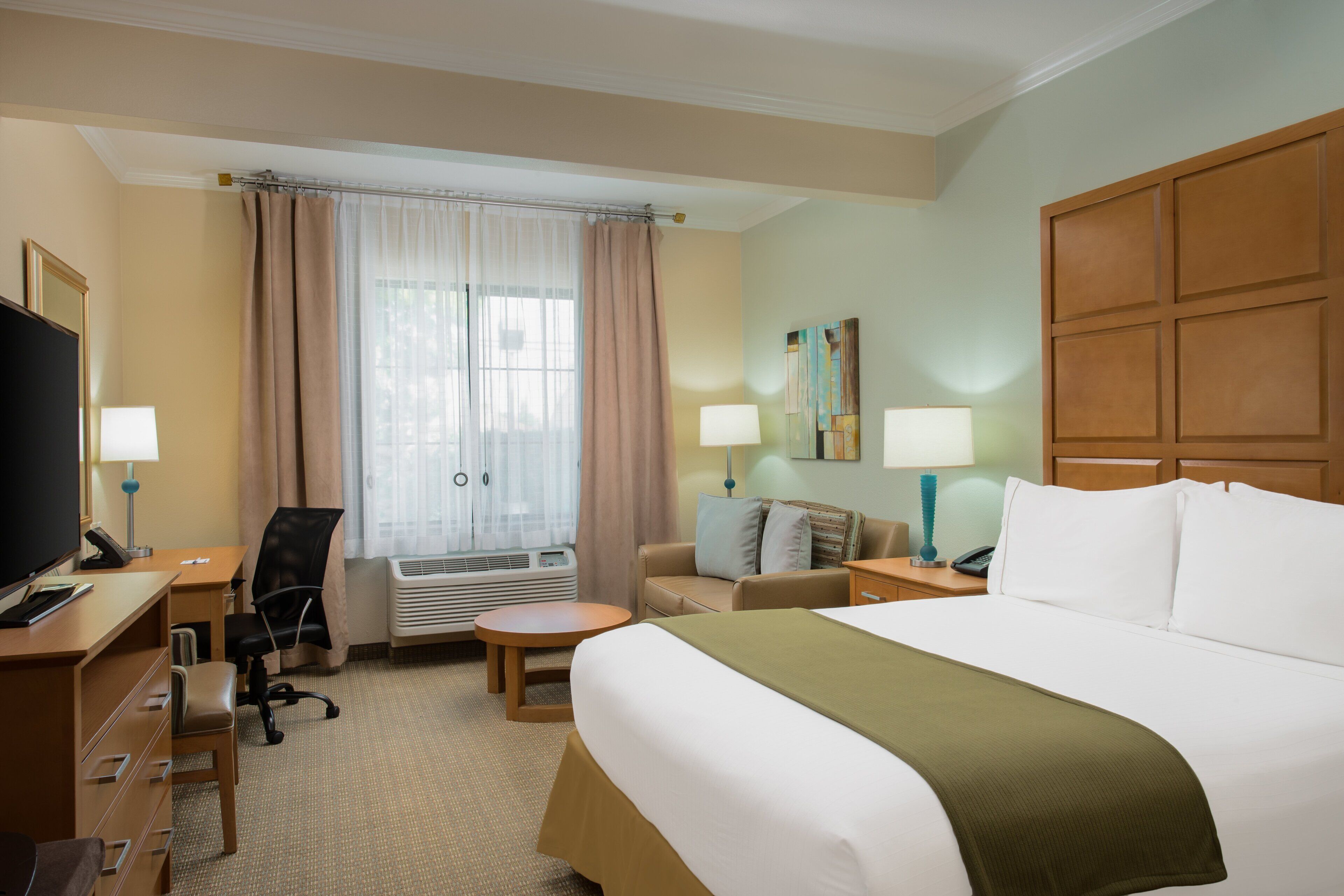 A hotel room with a bed, a desk with a computer, a chair, a small table, a lamp, a TV, and a window with curtains.