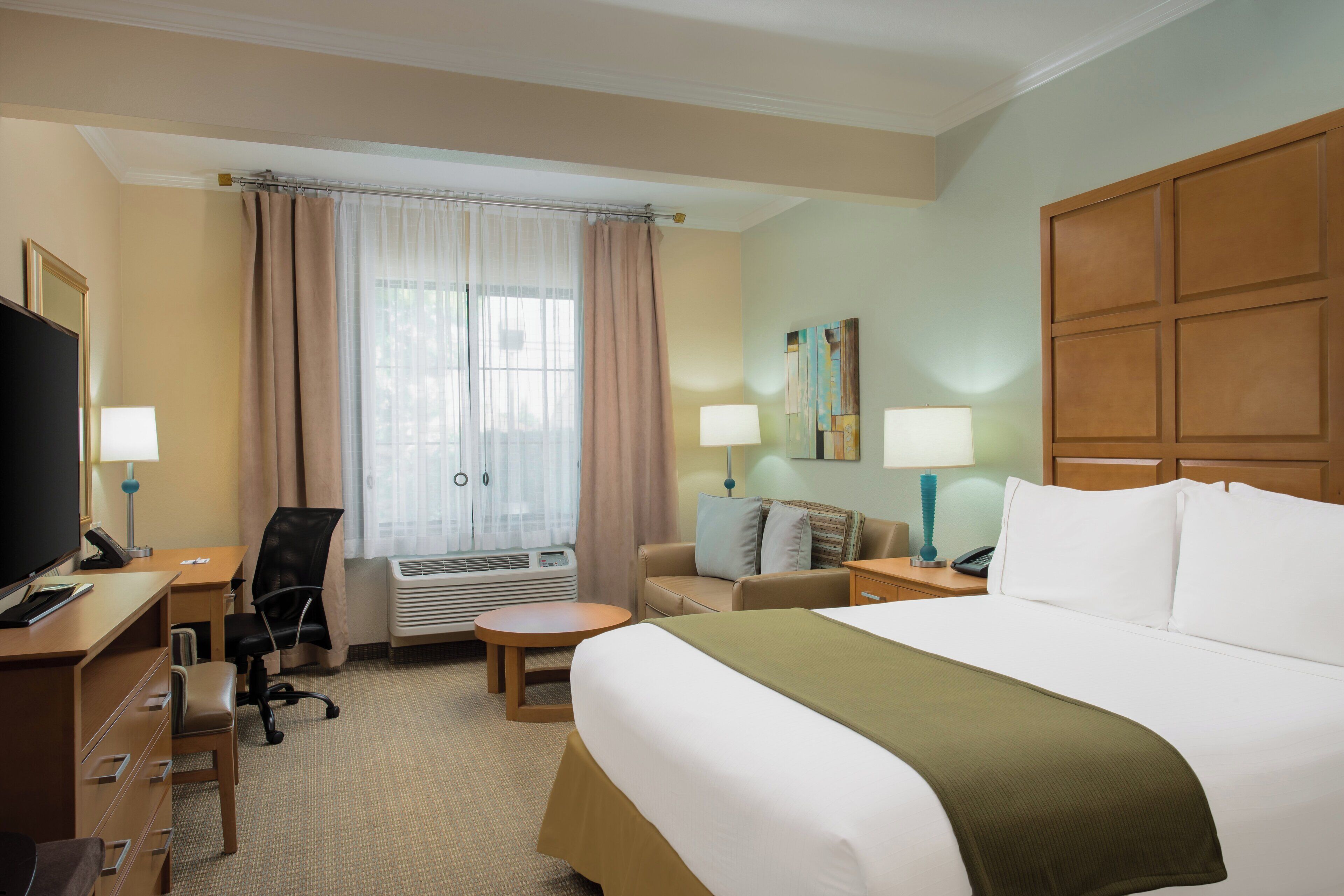 A hotel room with a bed, a desk with a computer, a chair, a small table, and a window with curtains.