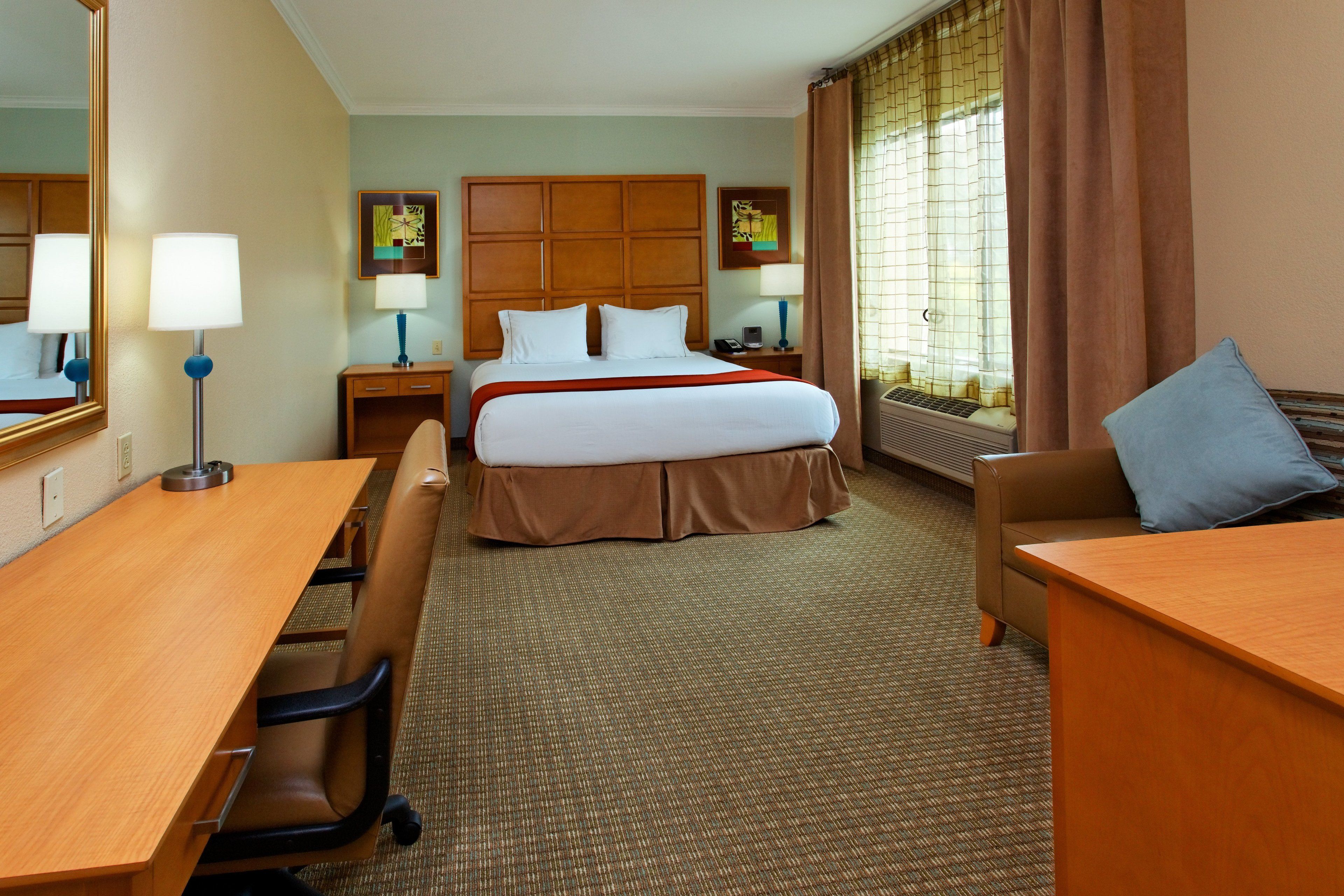 A hotel room with a large bed, a desk, a chair, a sofa, a nightstand, a lamp, and a window with curtains.
