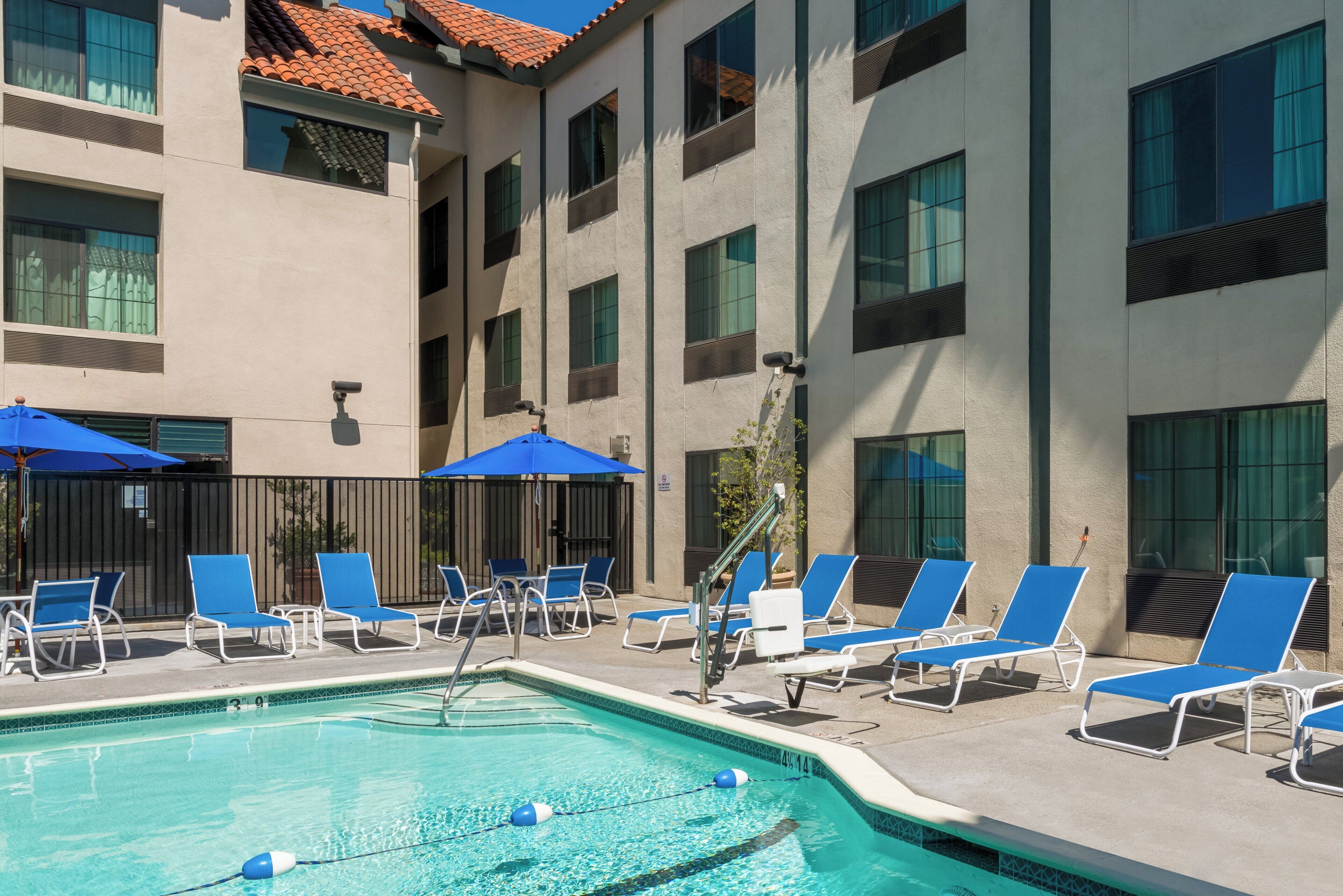 A swimming pool with blue lounge chairs and umbrellas, surrounded by a building with multiple windows and a tiled roof.