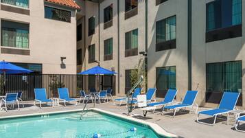 A swimming pool with blue lounge chairs and umbrellas, surrounded by a building with multiple windows and a tiled roof.