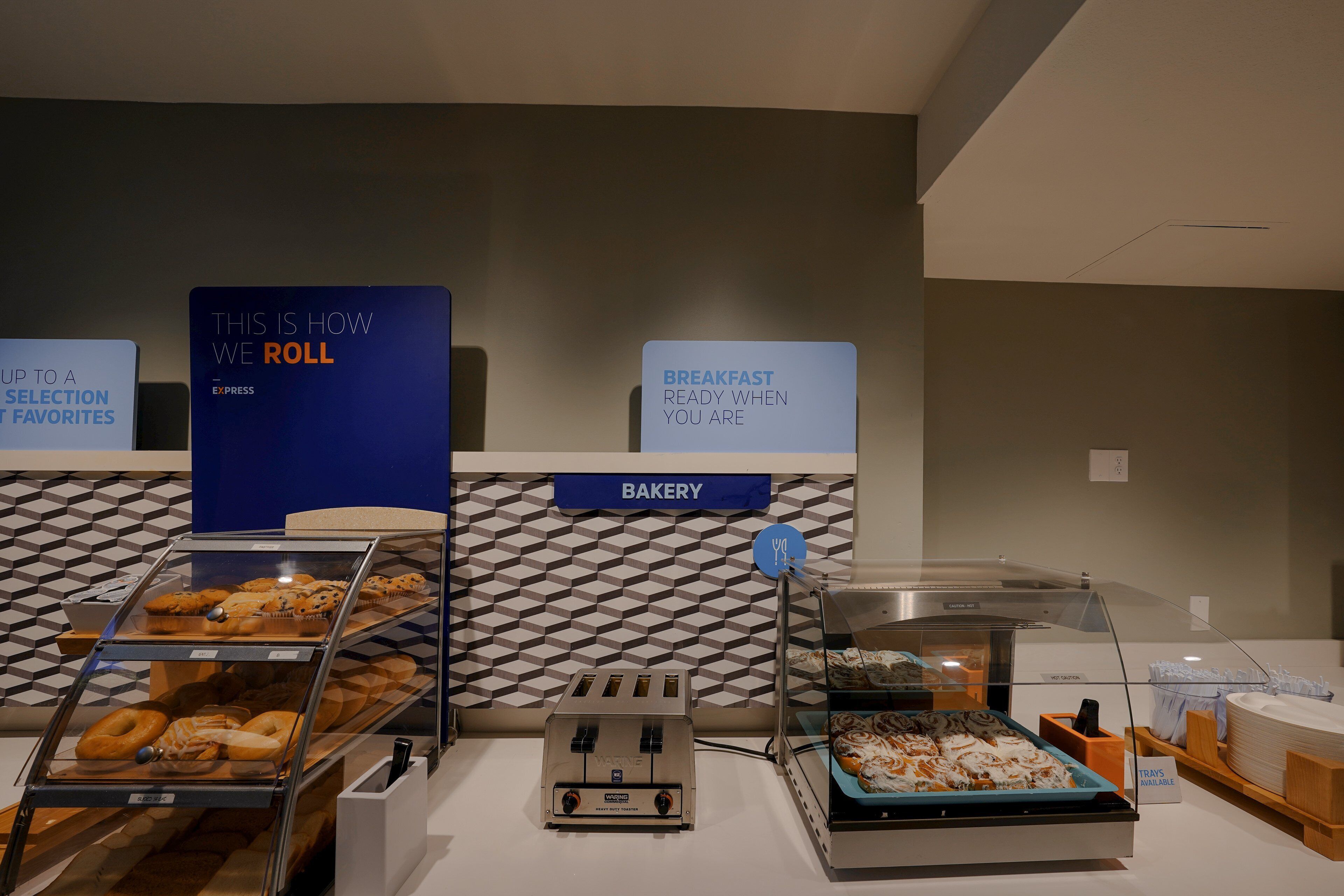 A bakery counter with various baked goods, a toaster, and a sign indicating breakfast is ready when you are.