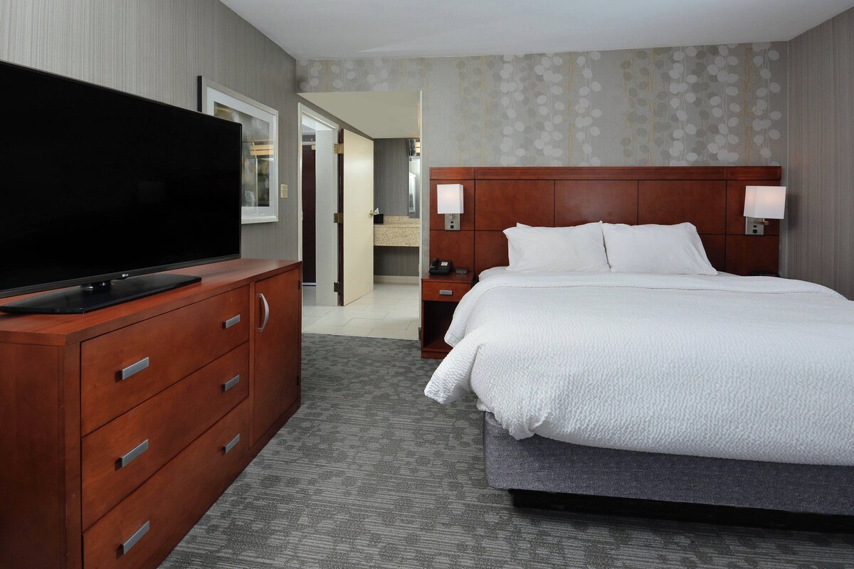 A hotel room with a large flat-screen TV on a wooden dresser, a bed with white linens, two bedside lamps, and a wooden headboard.