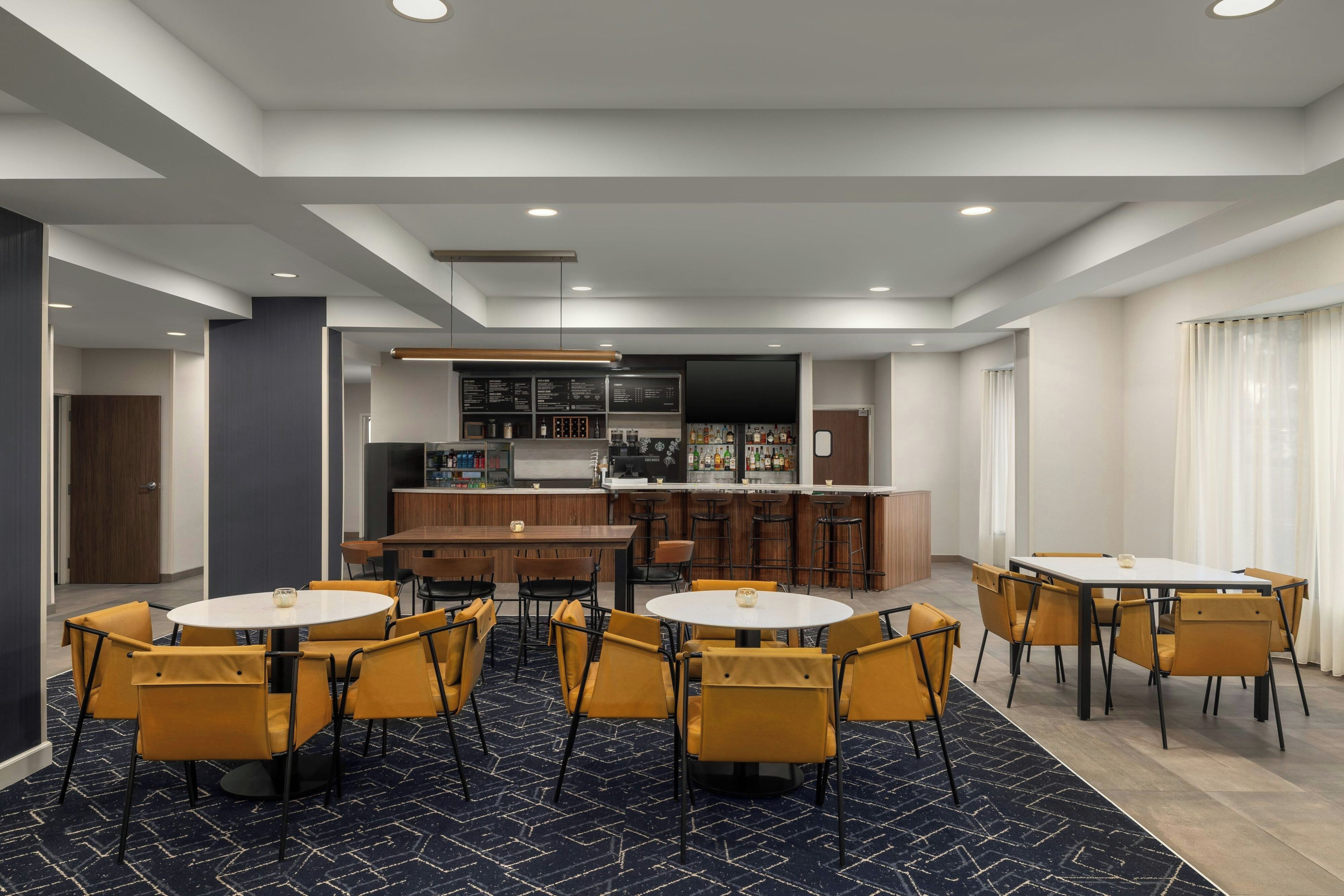 A modern hotel room with a bar area, multiple seating arrangements, and a large television screen.