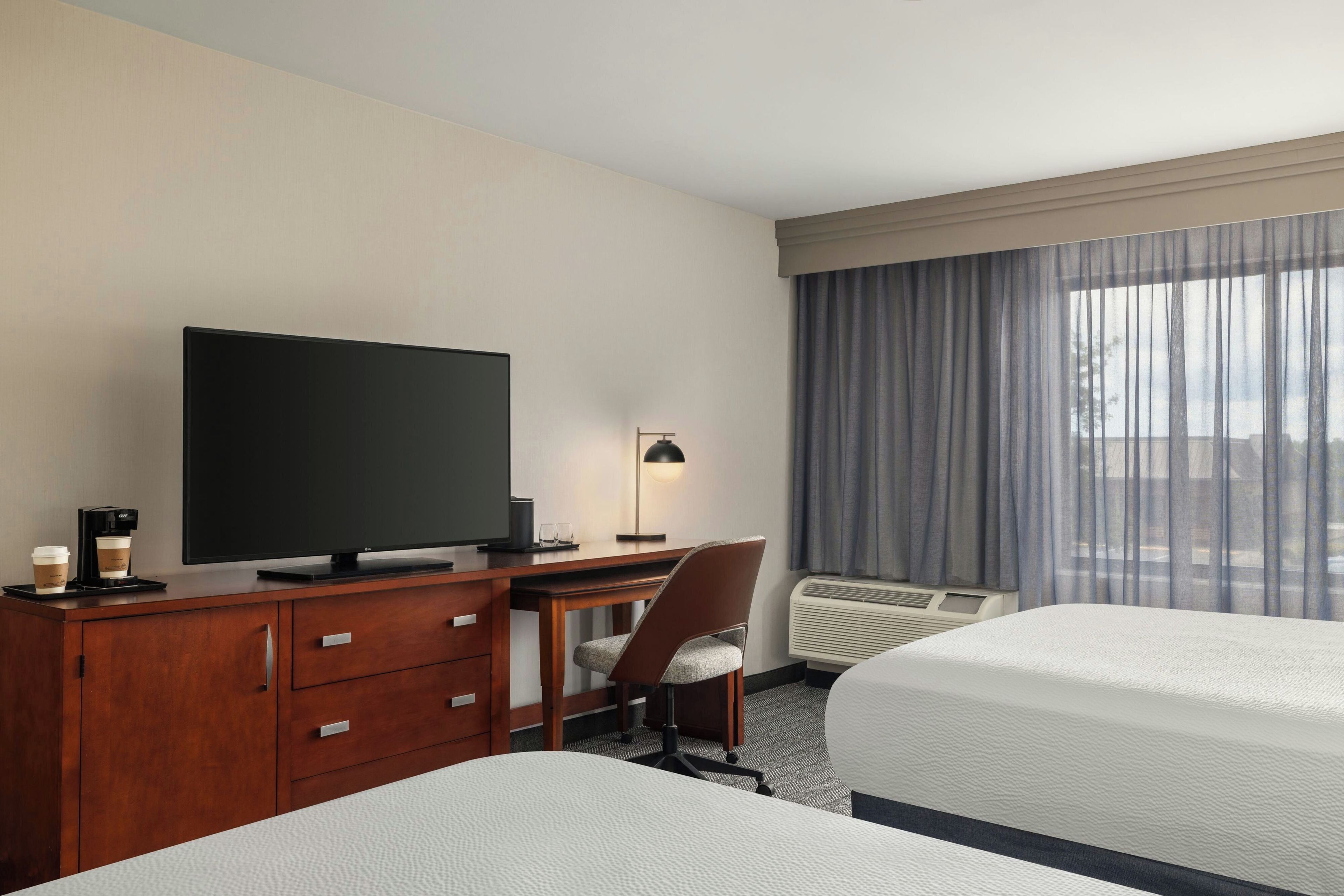 Habitación de hotel con televisión de pantalla plana, escritorio con cafetera, una silla, cama y ventana con cortinas.