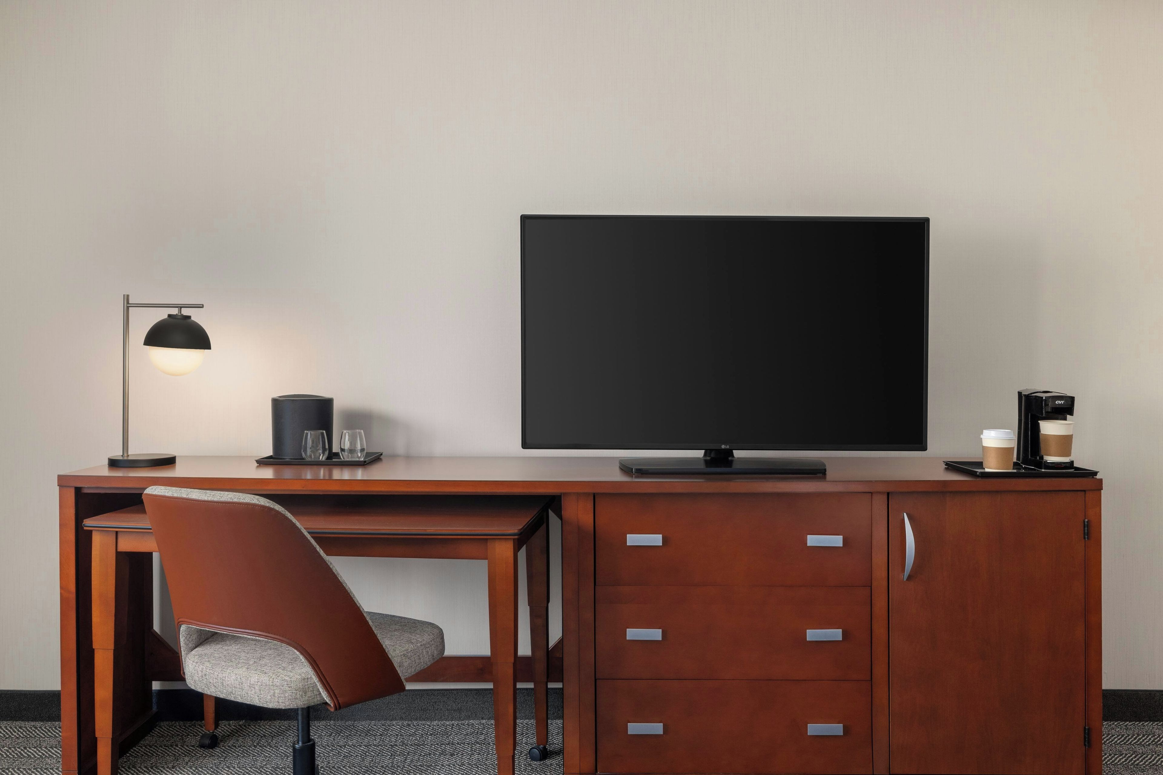 A hotel room with a wooden desk, a chair, a television, a coffee machine, and a cabinet with drawers.