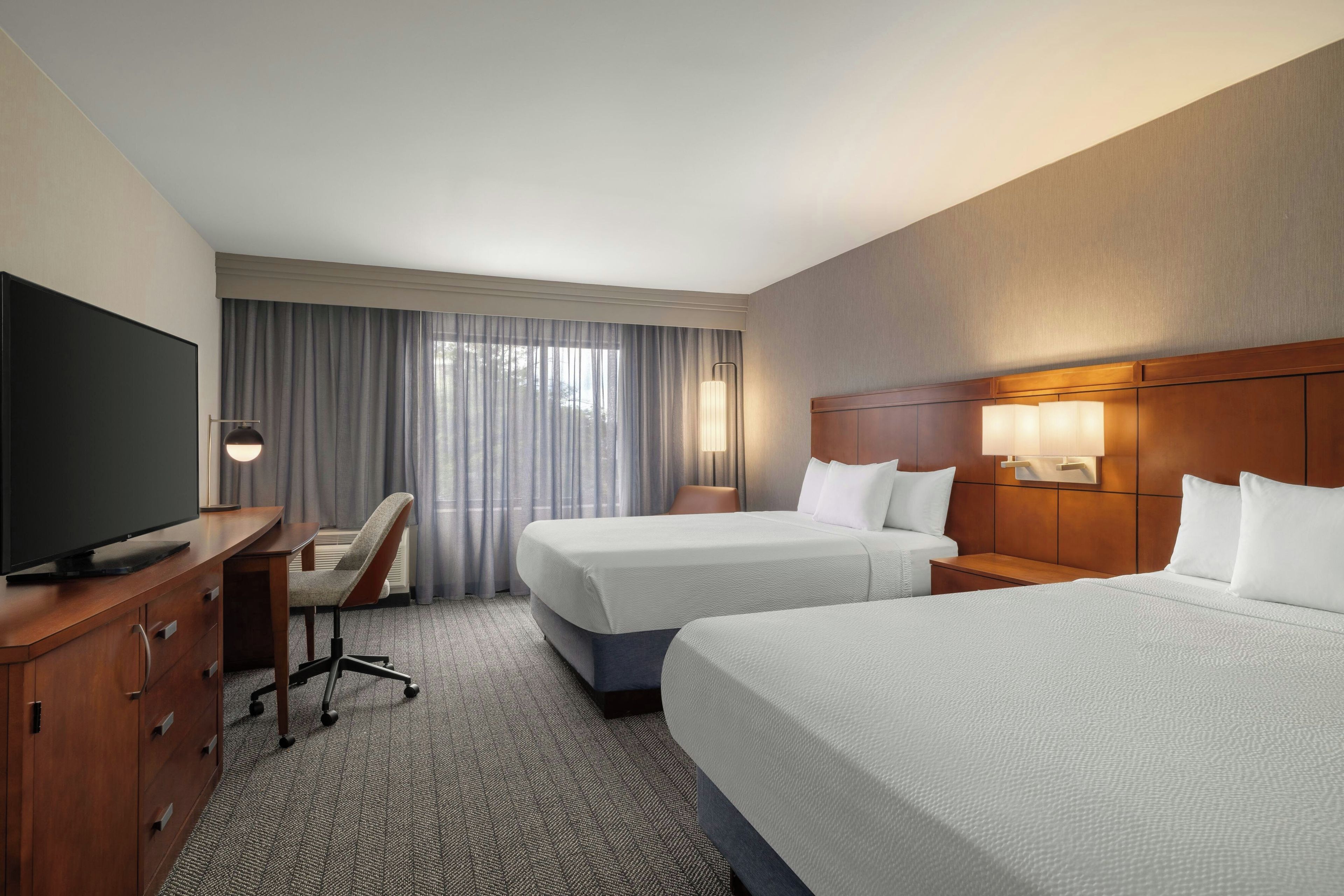 A hotel room with two beds, a desk with a TV, a chair, and a large window with curtains.