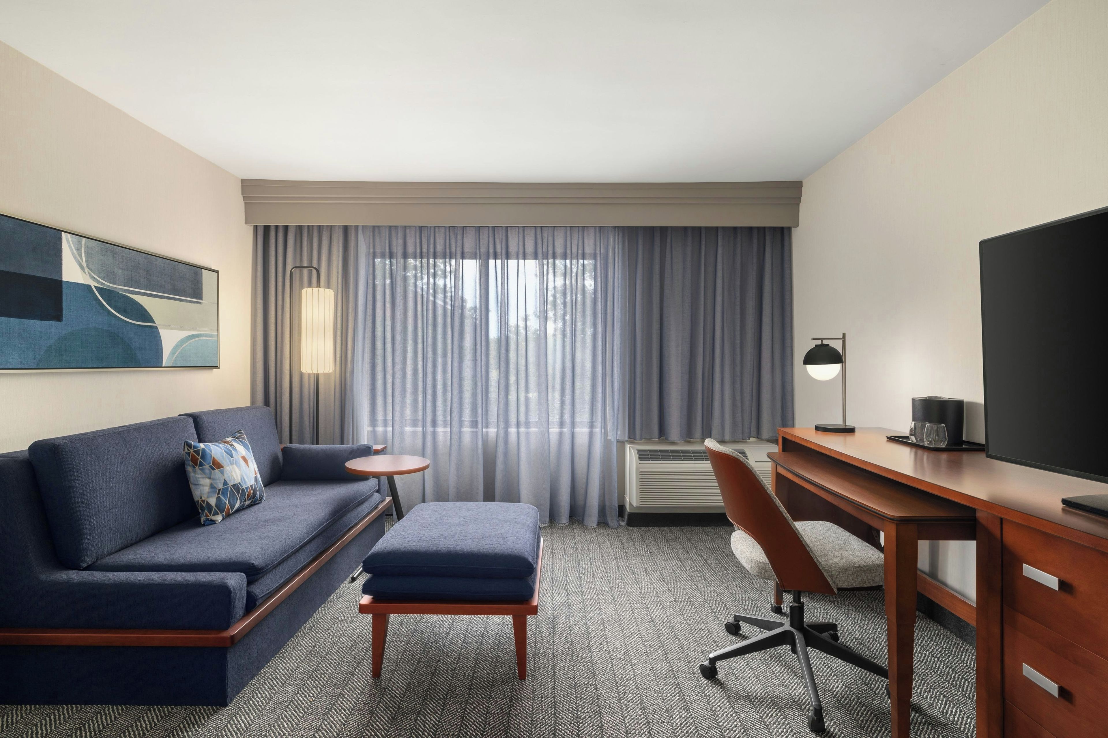 A hotel room with a sofa, a small table, a desk with a chair, a television, and a large window with curtains.
