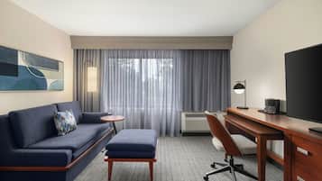 A hotel room with a sofa, a small table, a desk with a chair, a television, and a large window with curtains.
