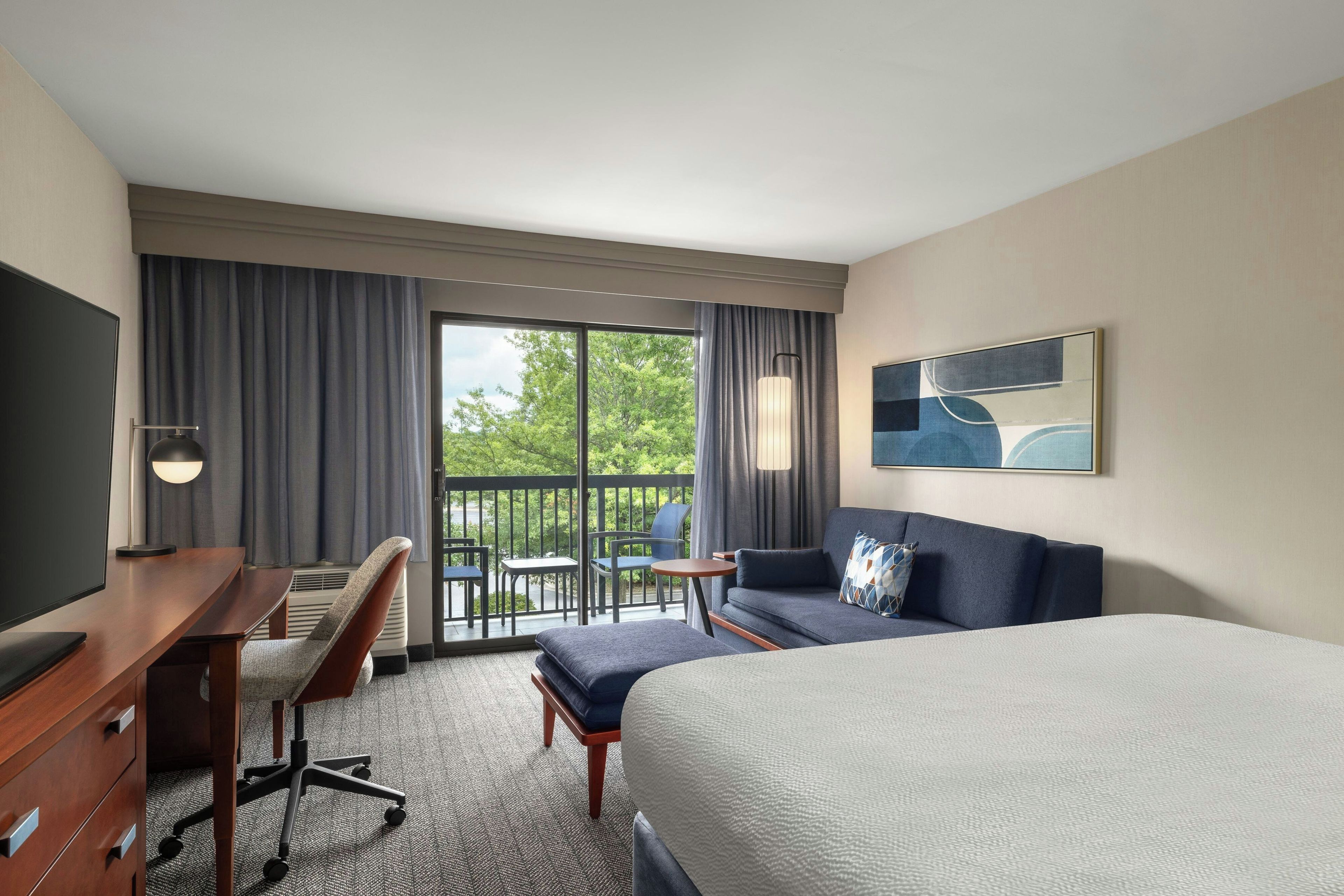Une chambre d’hôtel comprenant un lit, un bureau, une chaise, un canapé, une télévision, un tableau et un balcon avec vue sur les arbres.