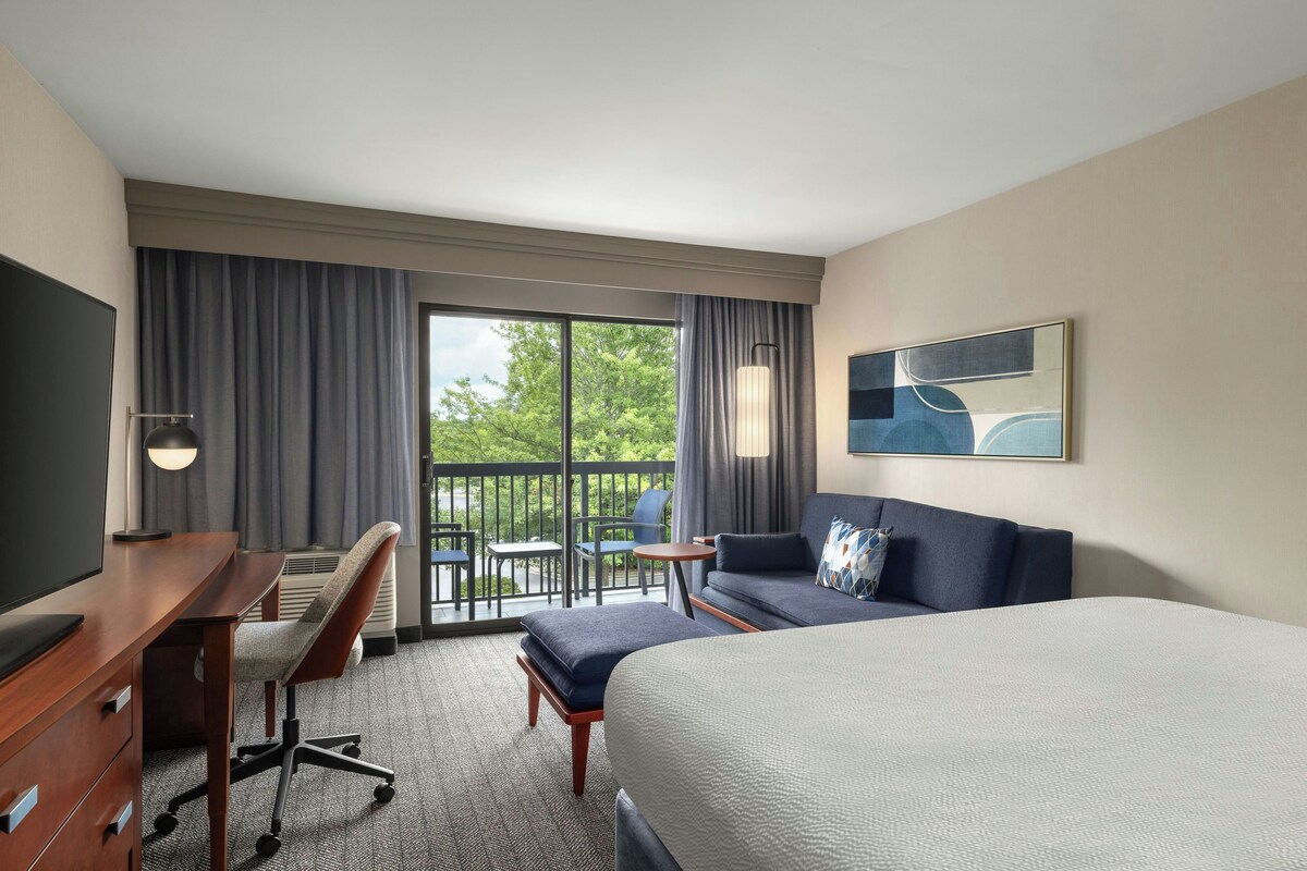 Une chambre d’hôtel comprenant un lit, un bureau, une chaise, un canapé, une télévision, un tableau et un balcon avec vue sur les arbres.