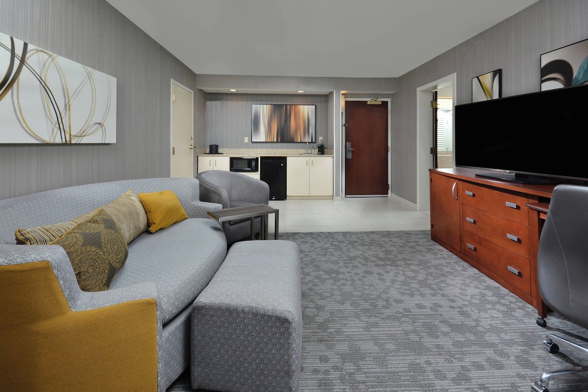 A modern hotel room with a grey sofa, a small table, a television, a wooden cabinet, and a kitchenette in the background.