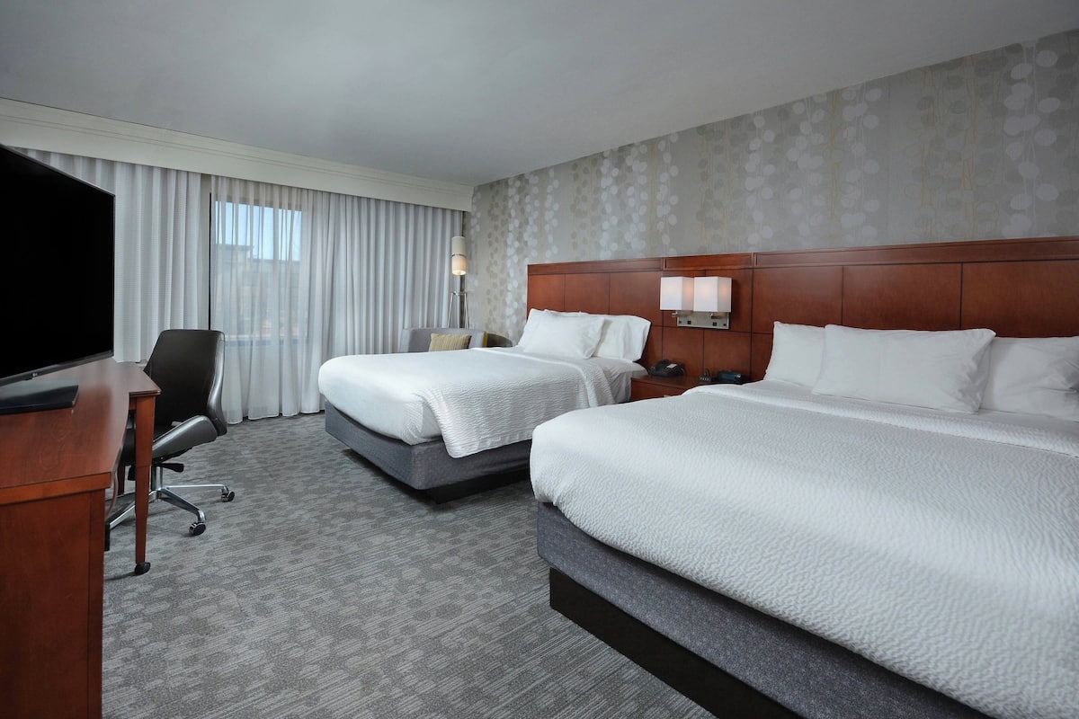 A hotel room with two beds, a desk, a TV, and a large window with curtains.