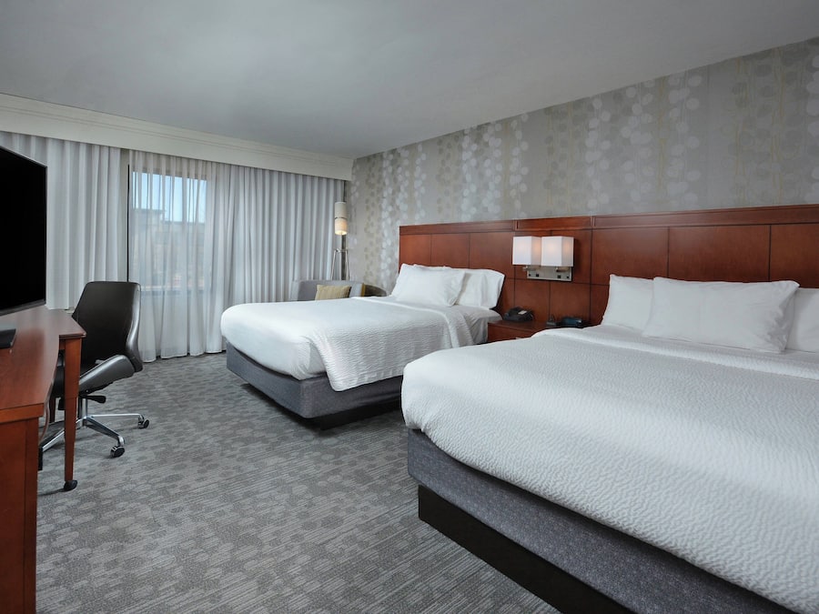A hotel room with two beds, a desk, a TV, and a large window with curtains.