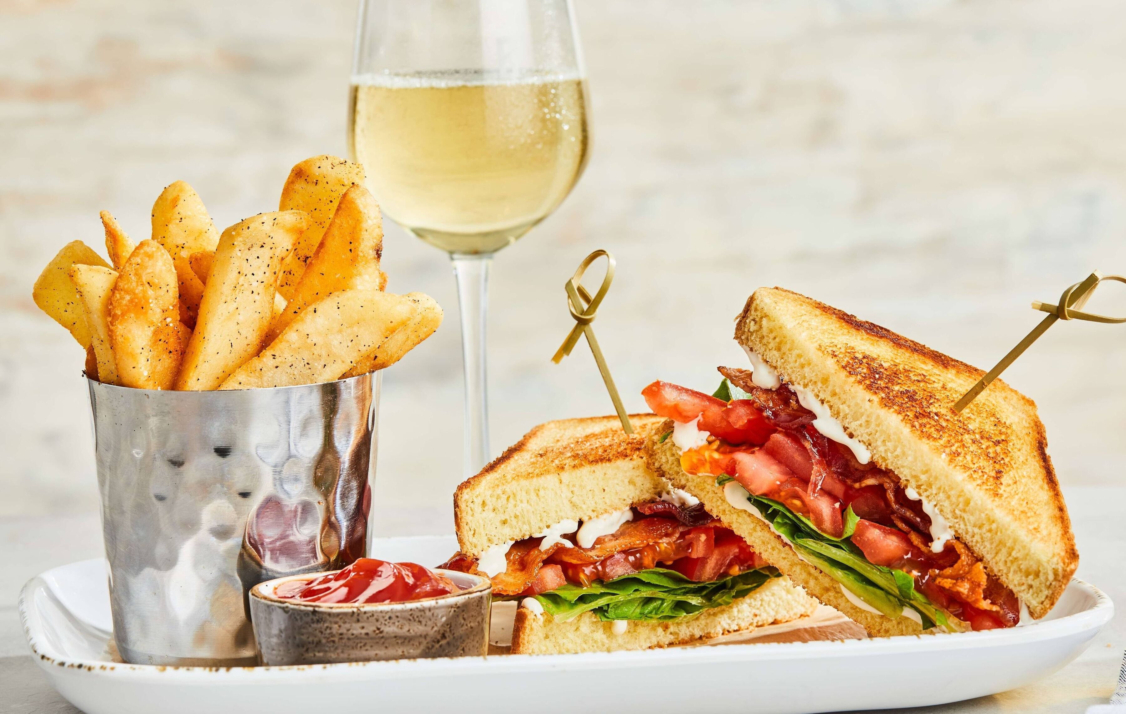 A plate with a grilled sandwich, a glass of white wine, and a container of fries.