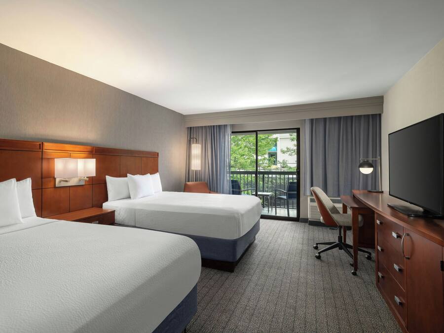 A hotel room with two beds, a desk, a television, and a balcony with outdoor furniture.