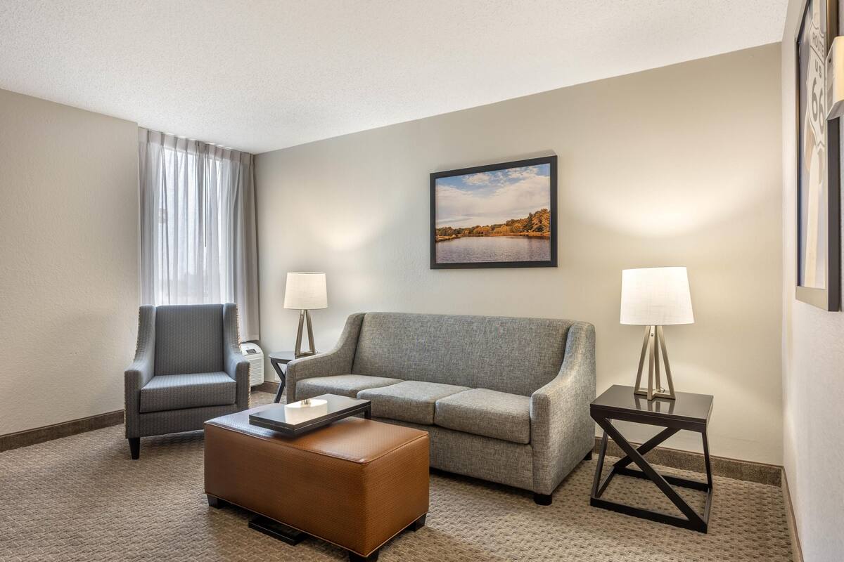 A hotel room with a grey sofa, a brown ottoman, two side tables with lamps, a chair, and a framed picture on the wall.