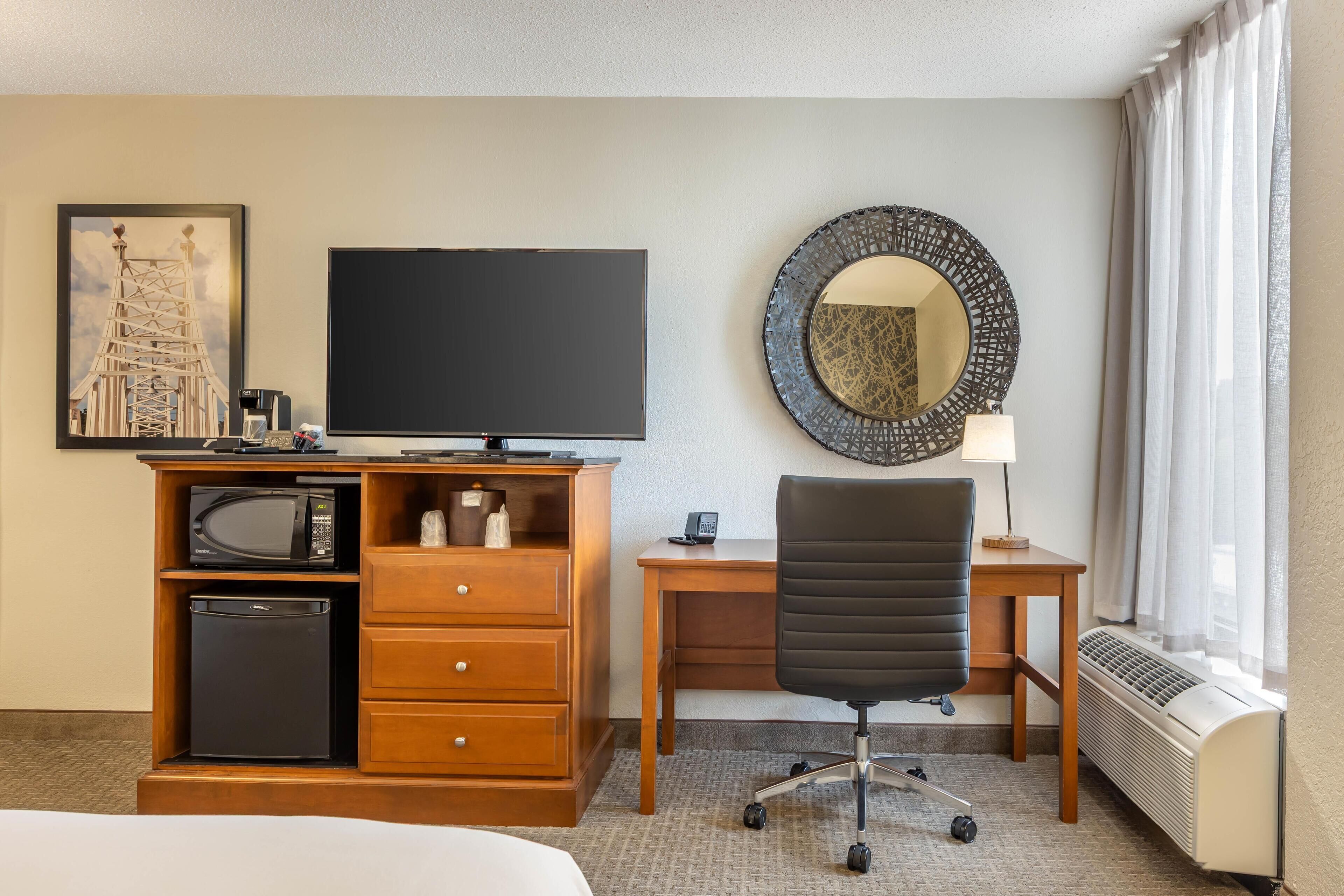 A hotel room with a bed, a desk with a computer, a chair, a microwave, a small refrigerator, a flat-screen TV, a decorative mirror, and a window with curtains.
